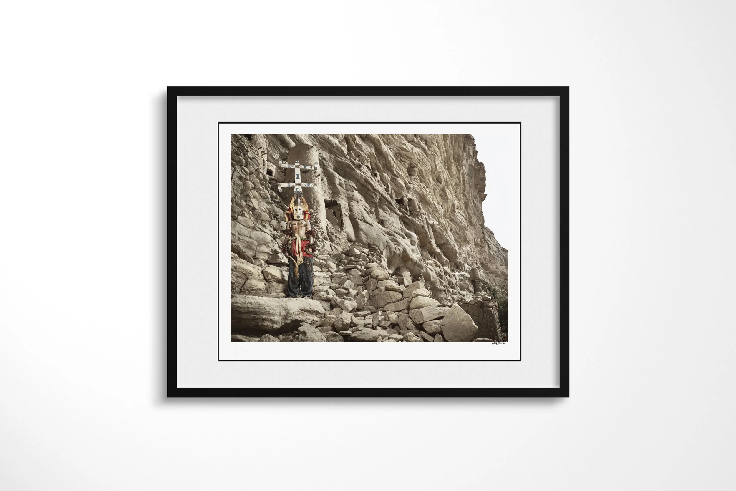 Framed presentation of Kanaga (Mask) in a black frame and white mount, showing a Dogon Kanaga mask with a tall double-cross form standing below the Bandiagara cliffs.