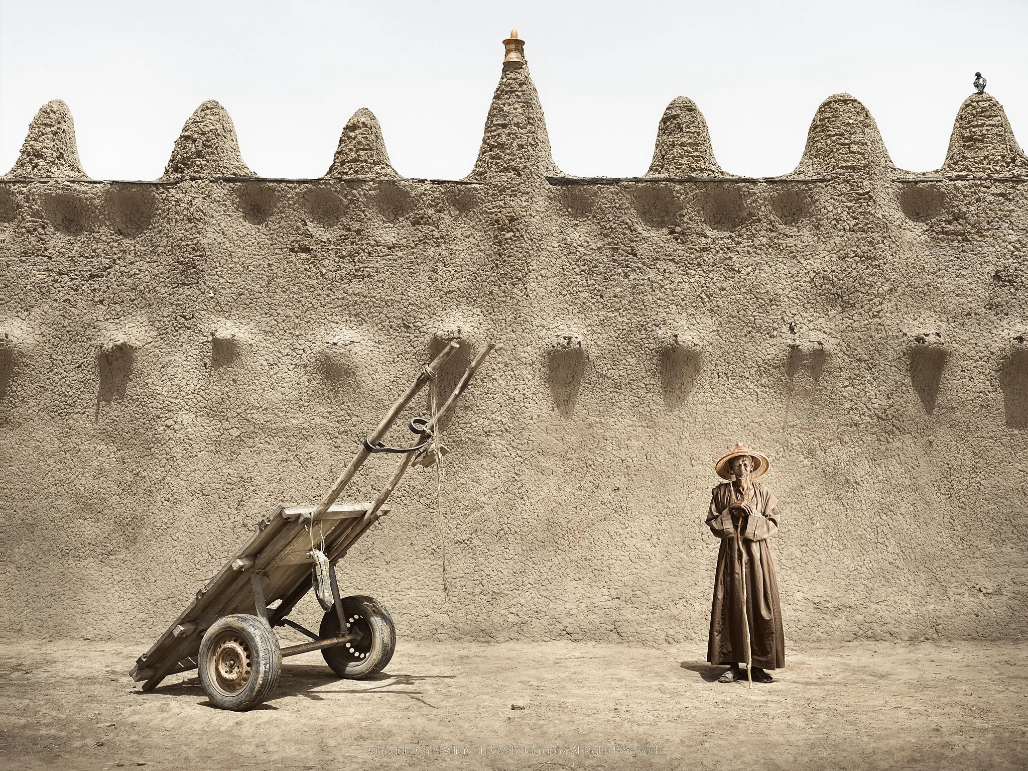 Ouman standing beside a two-wheeled handcart against the sculpted wall of an earthen mosque.