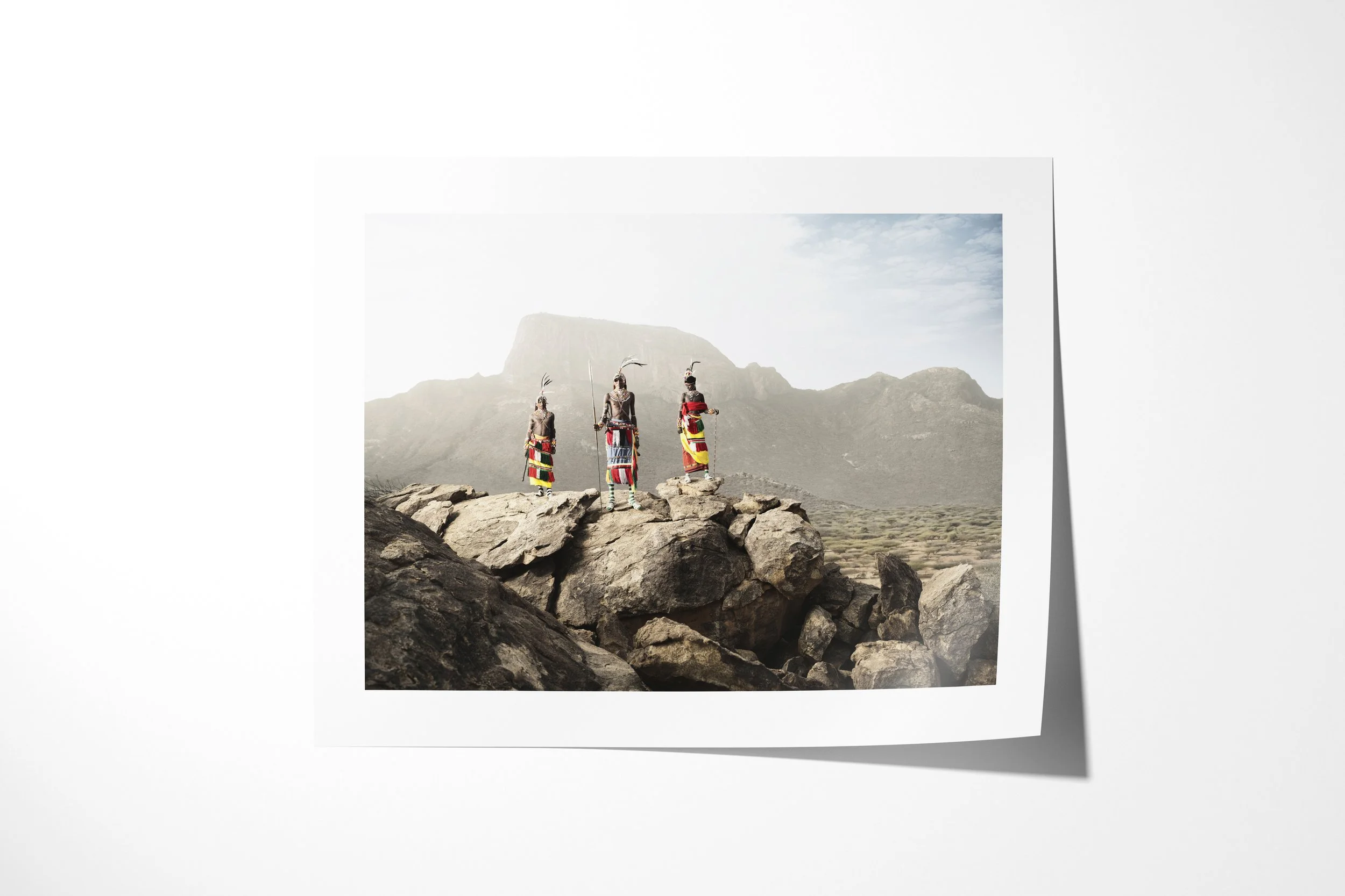 Fine art paper print of “The Warriors of Mt.Poi”, showing three Samburu warriors in bright dress and feathered headdresses standing on rocks before Mount Poi.