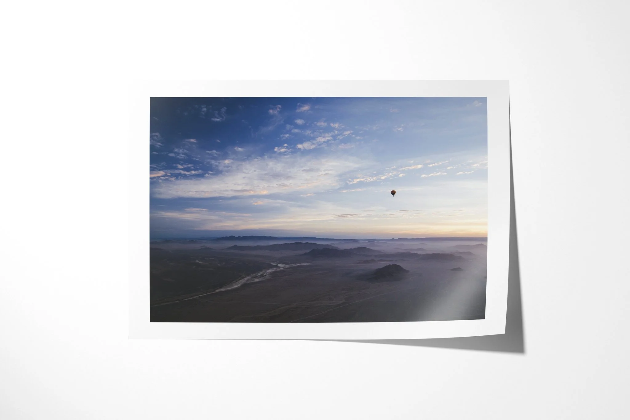 Fine art paper print of Over The Dunes, showing small hot-air balloon above an expansive arid landscape of ridges, valleys and pale winding channels at dawn.