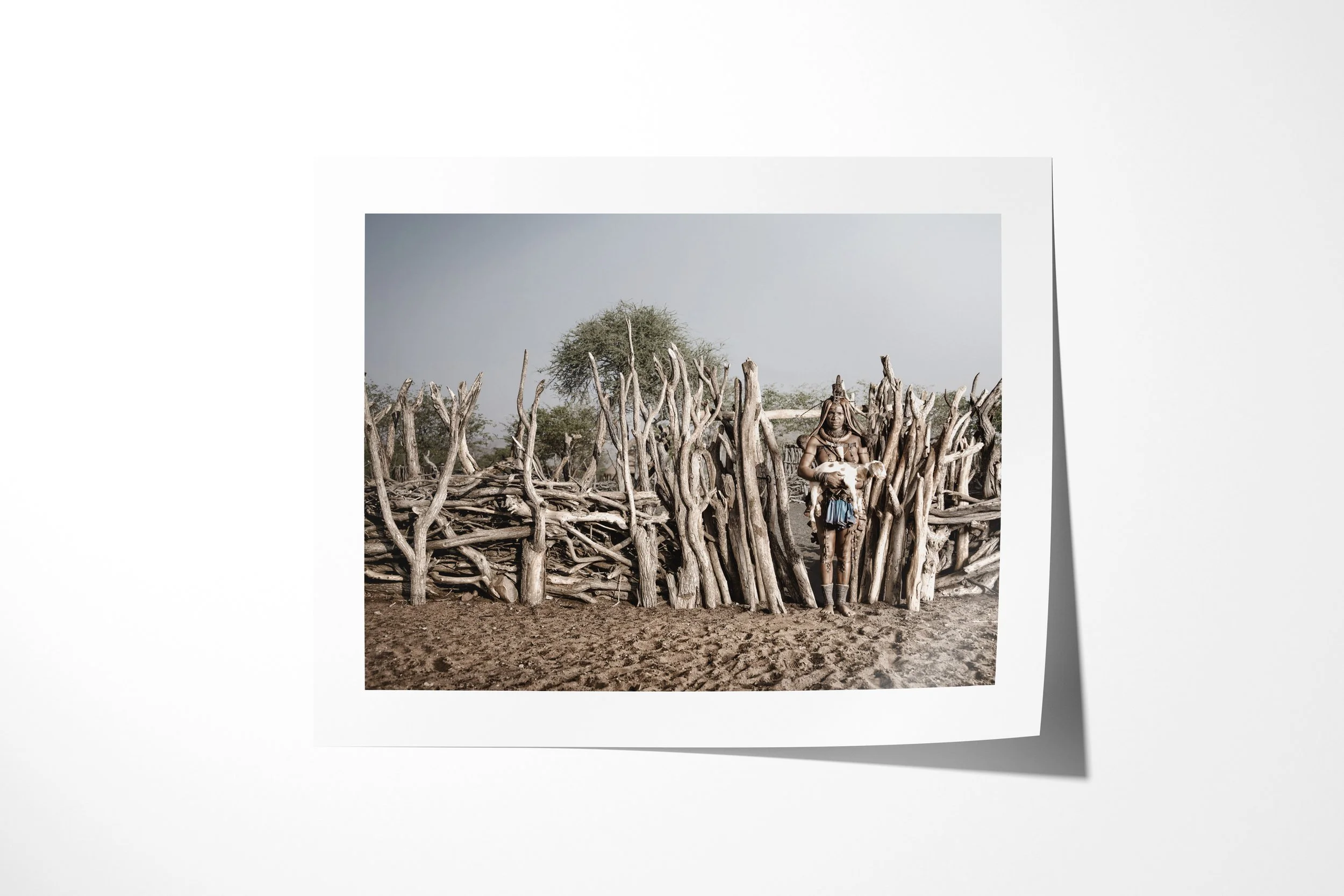 Fine art paper print of Twahenwa III, showing Twahenwa holding a young goat beside the branch enclosure of a Himba homestead.