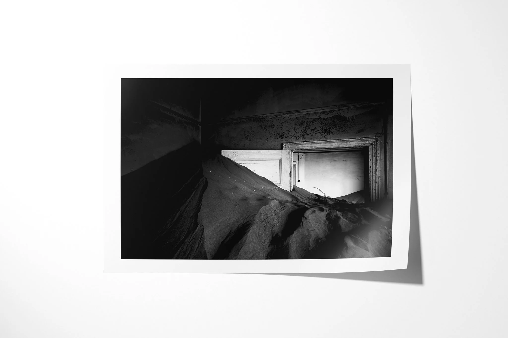 Fine art paper print of Doorway I, showing a steep sand drift filling a dark abandoned room and rising through a partly open doorway in black and white.