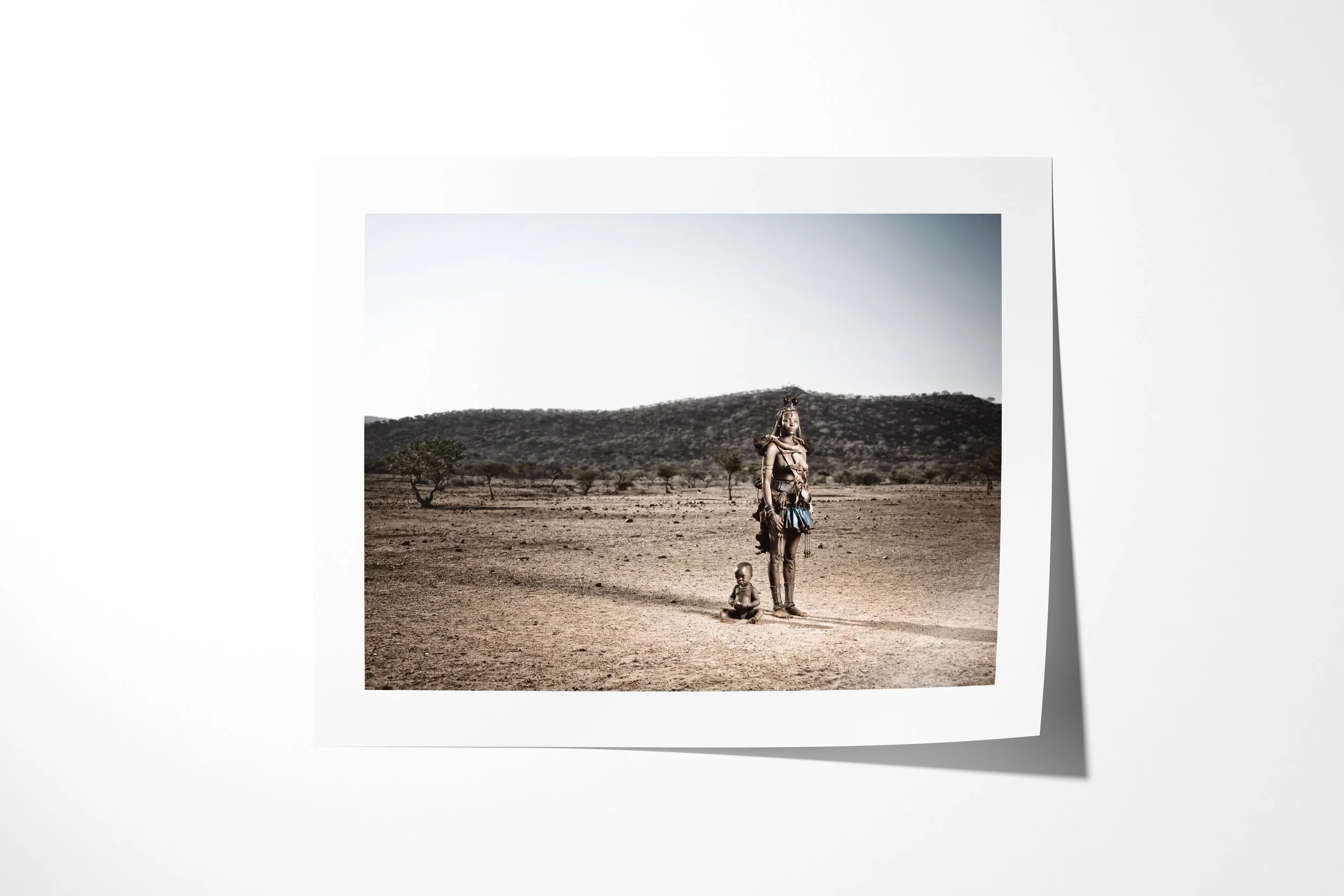 Fine art paper print of Mburaivaka & Her Child, showing Mburaivaka standing with her child in an expansive dry landscape backed by low hills.