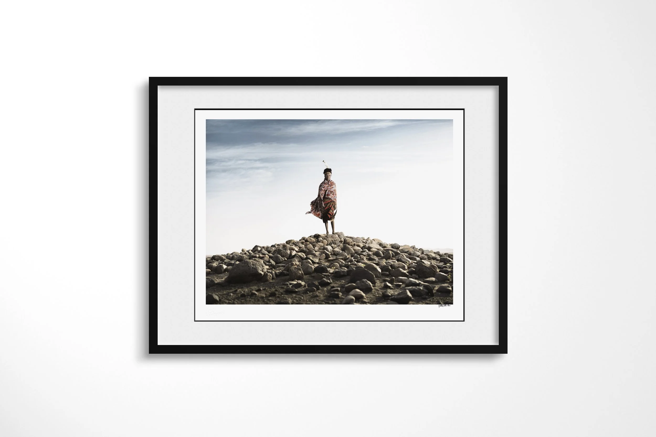 Framed presentation of Lomus - “The King” in a black frame and white mount, showing Lomus standing on a mound of rounded rocks as wind lifts his patterned cloth.