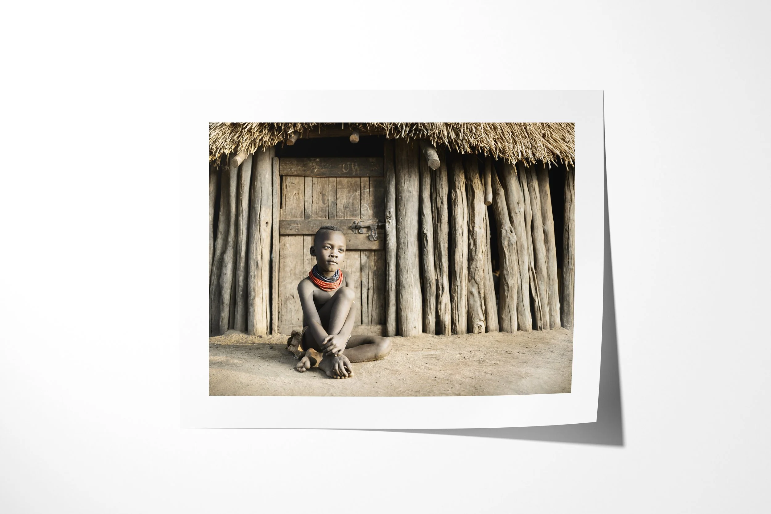 Fine art paper print of Kayo, showing a Kara child seated before the timber doorway of a thatched family house.