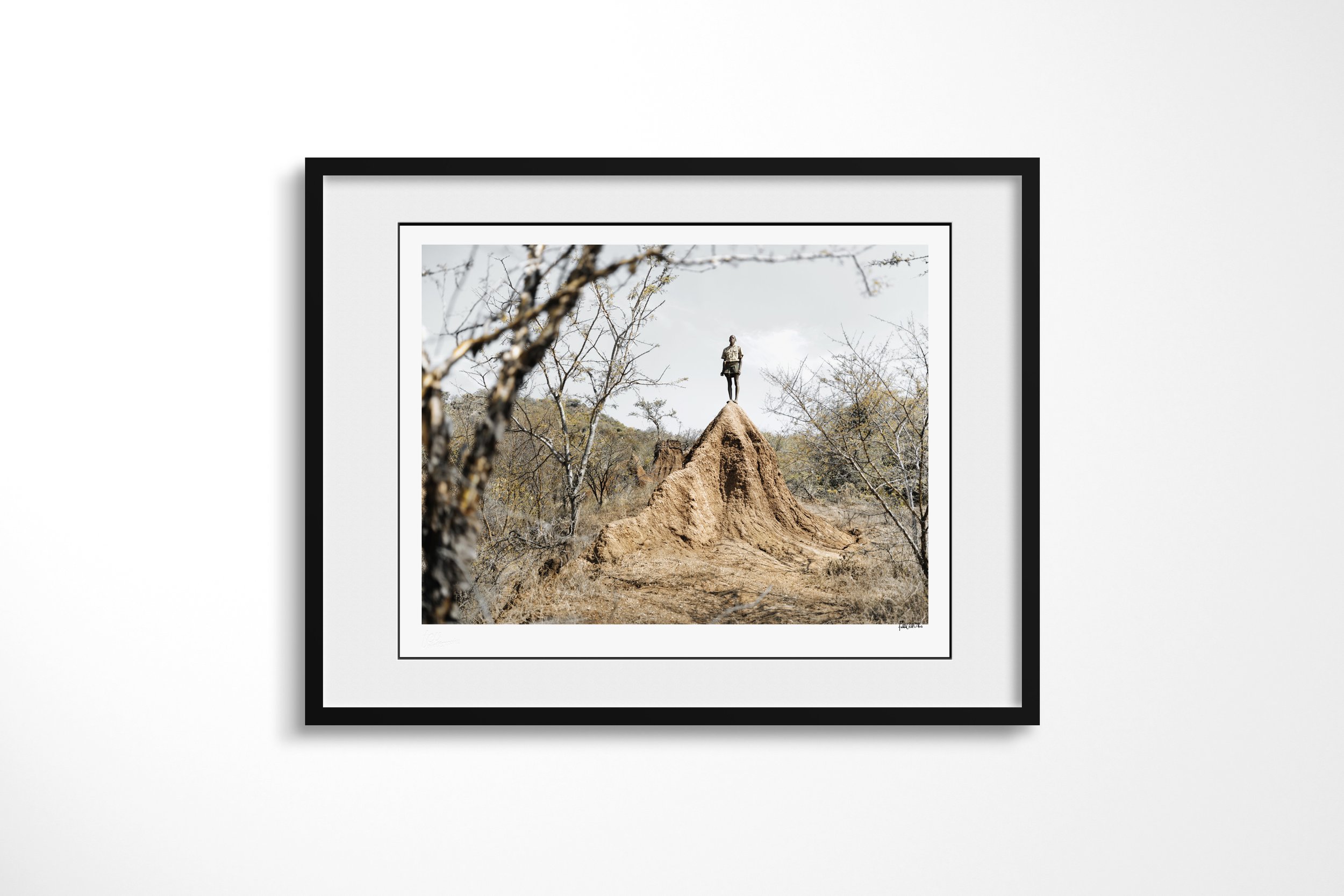 Framed presentation of Dheta in a black frame and white mount, showing a Banna figure standing on the peak of a large termite mound in dry woodland.