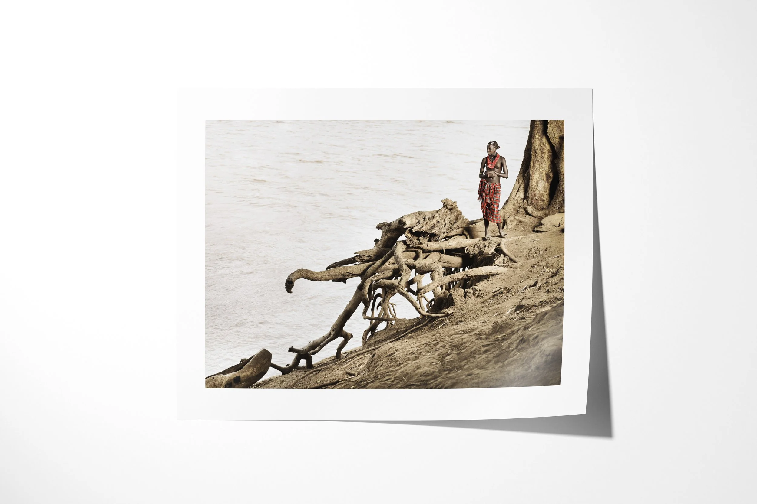Fine art paper print of Yierar, showing a Daasanach man standing beside exposed tree roots on the bank of the Omo River.