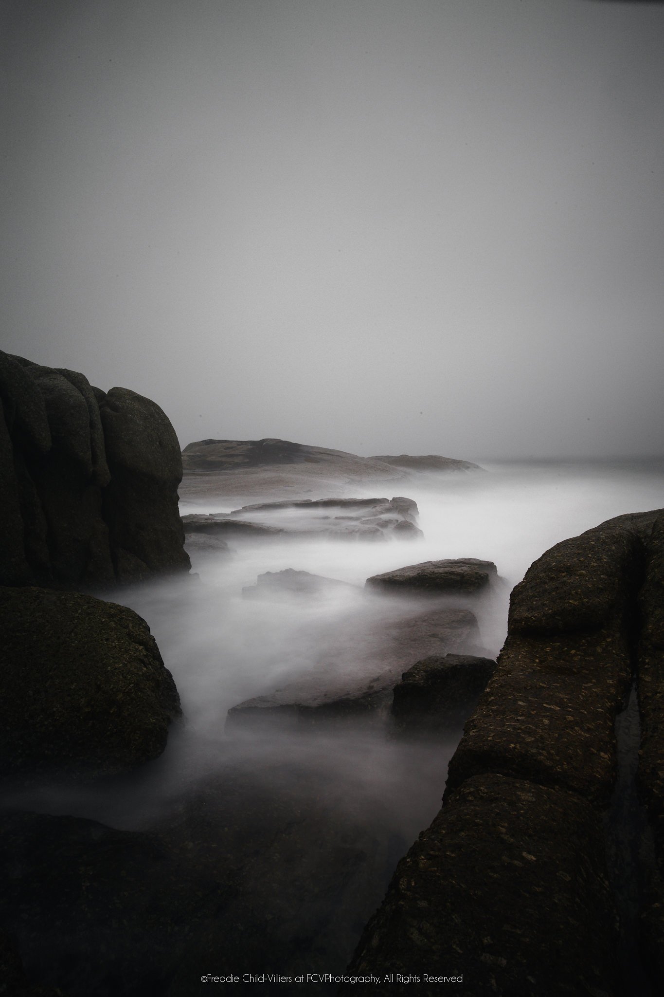 Dark coastal boulders framing pale long-exposure seawater beneath a blank grey mist.