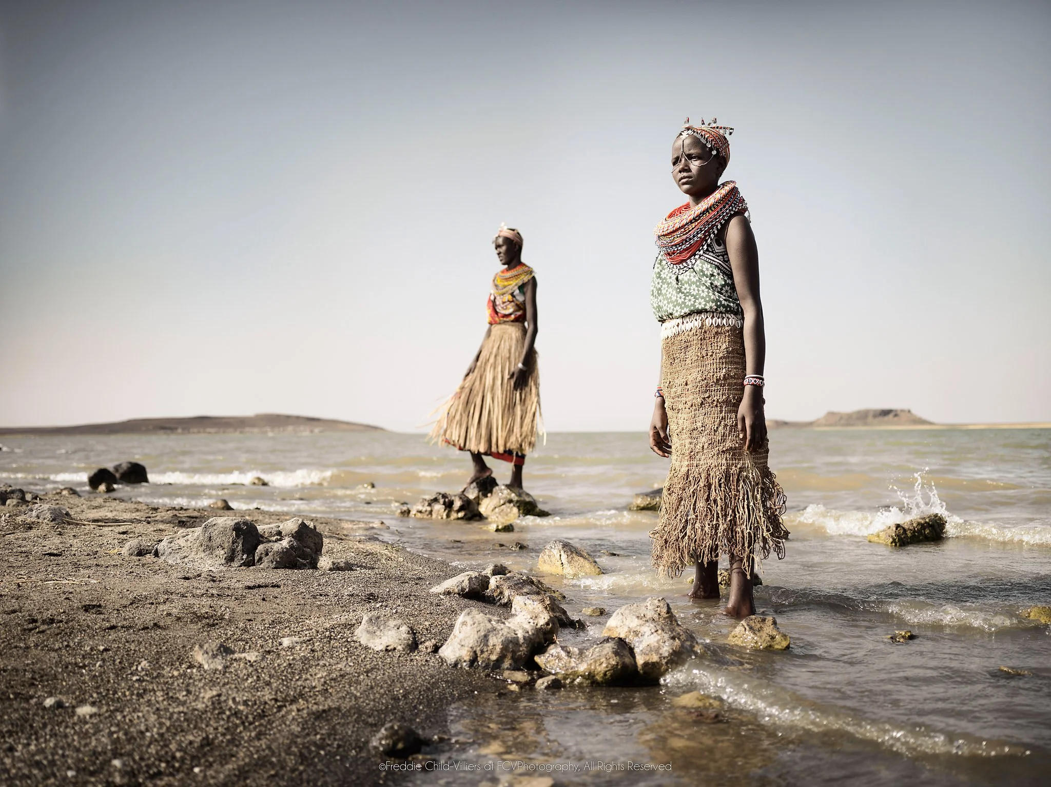 Lawra & Nadirie, two El Molo women in fibre garments standing on the rocky shore of Lake Turkana.