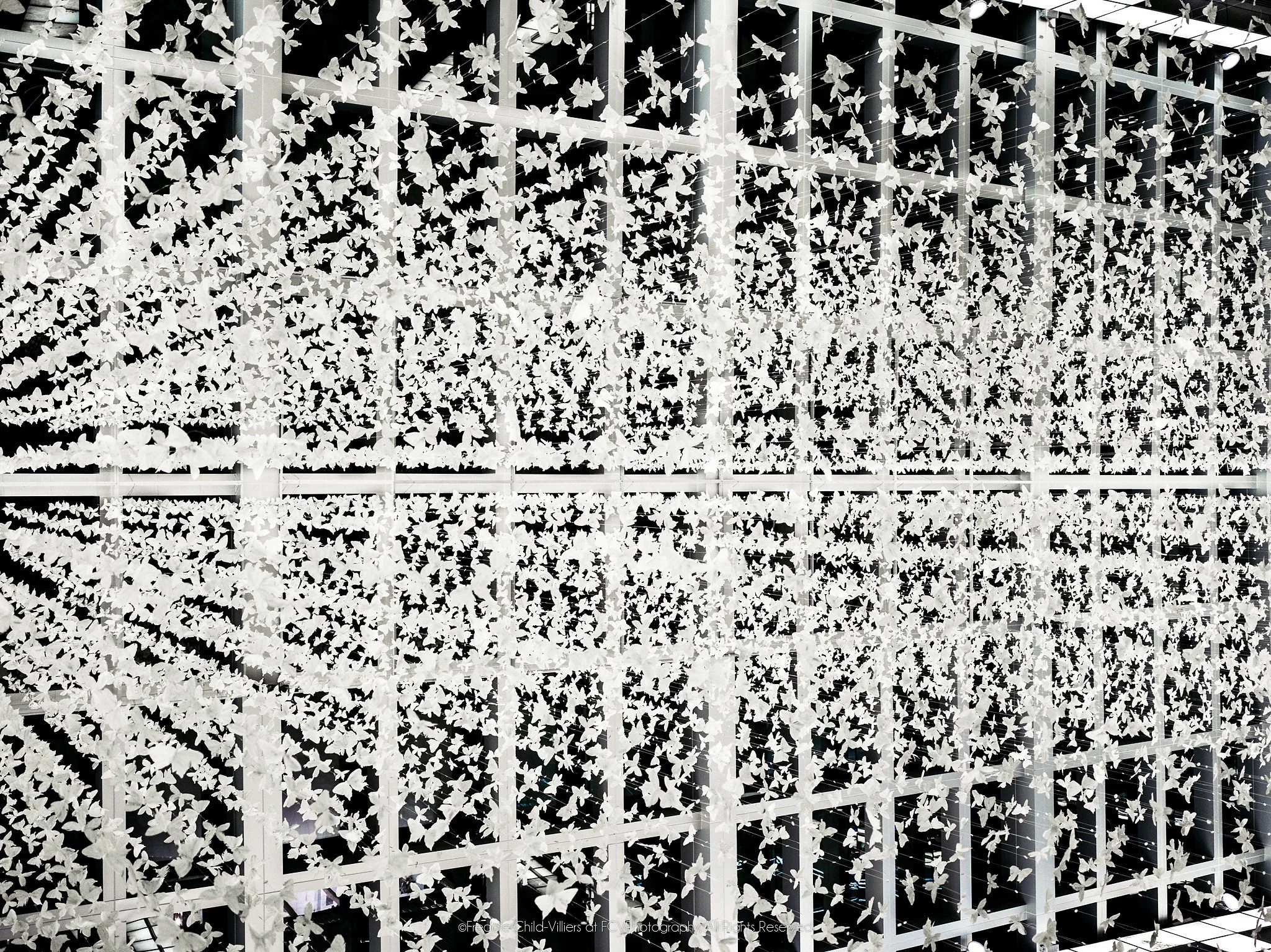 Hundreds of pale butterfly-shaped elements suspended on wires across a dark architectural grid.