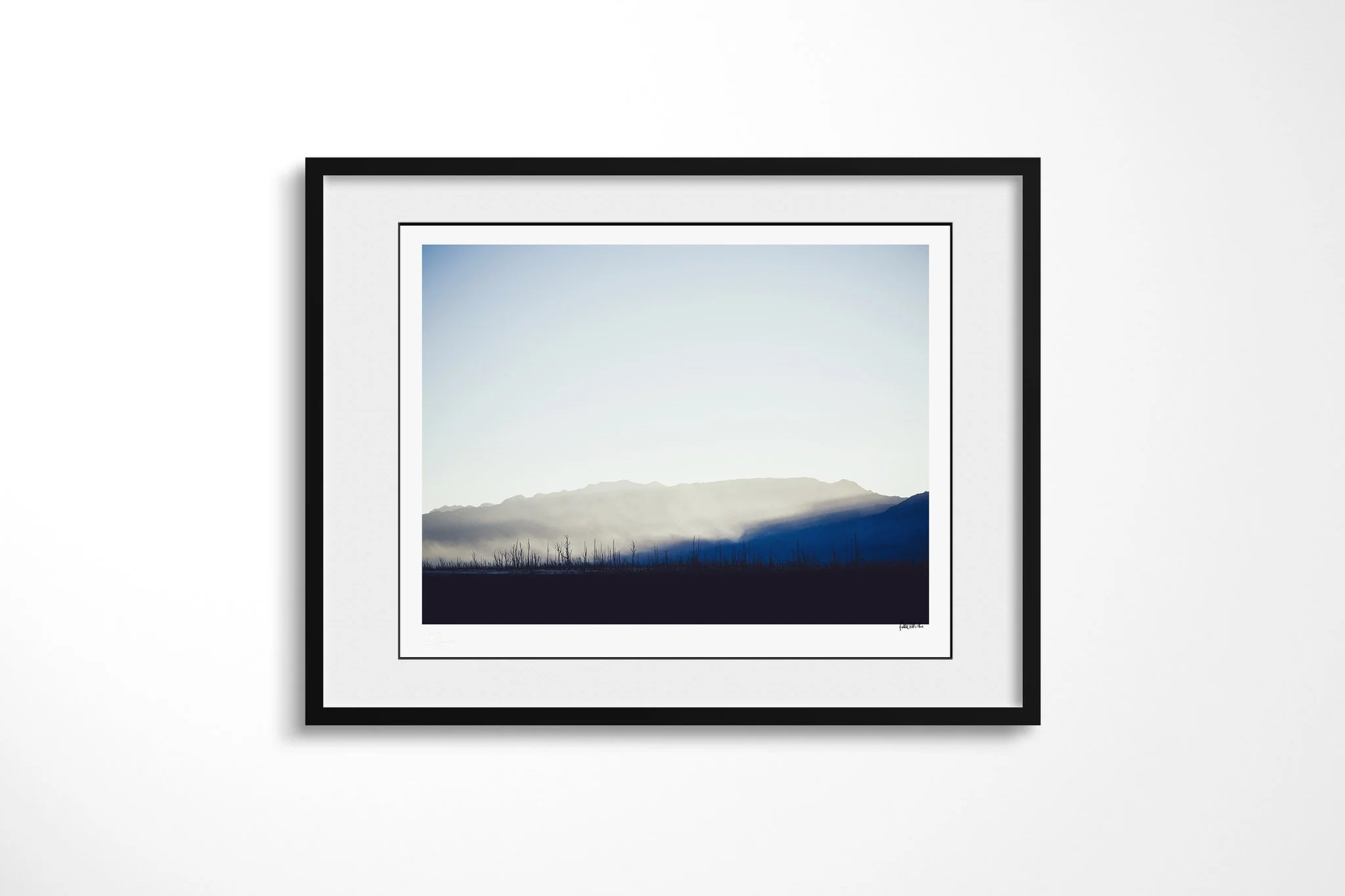 Framed fine art presentation of Drought in a black frame with a white mount, depicting bare blackened trees below a mountain ridge, with pale mist lit above a deep blue shadow.