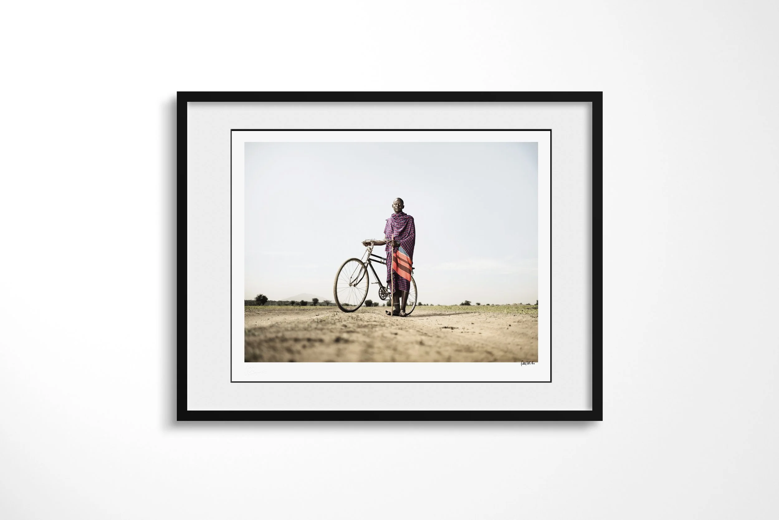 Framed presentation of Kimerei in a black frame and white mount, showing a Maasai man standing beside his bicycle on an open dirt road.