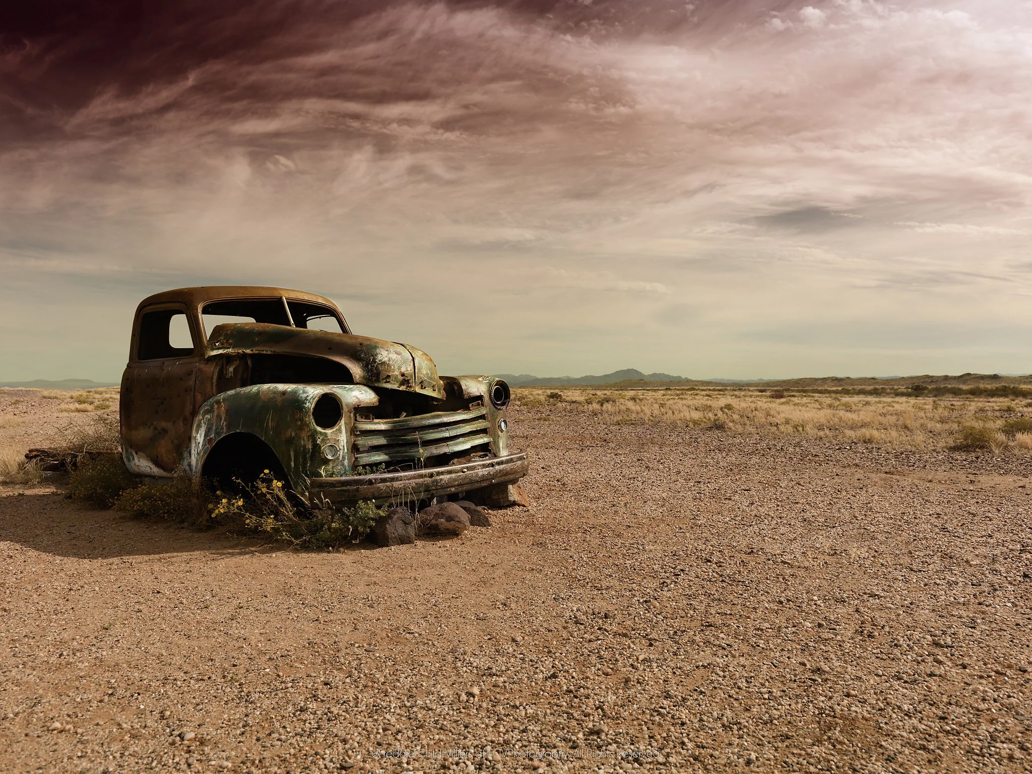 Rusted vintage vehicle abandoned on a stony arid plain beneath dark, textured clouds and distant low mountains.
