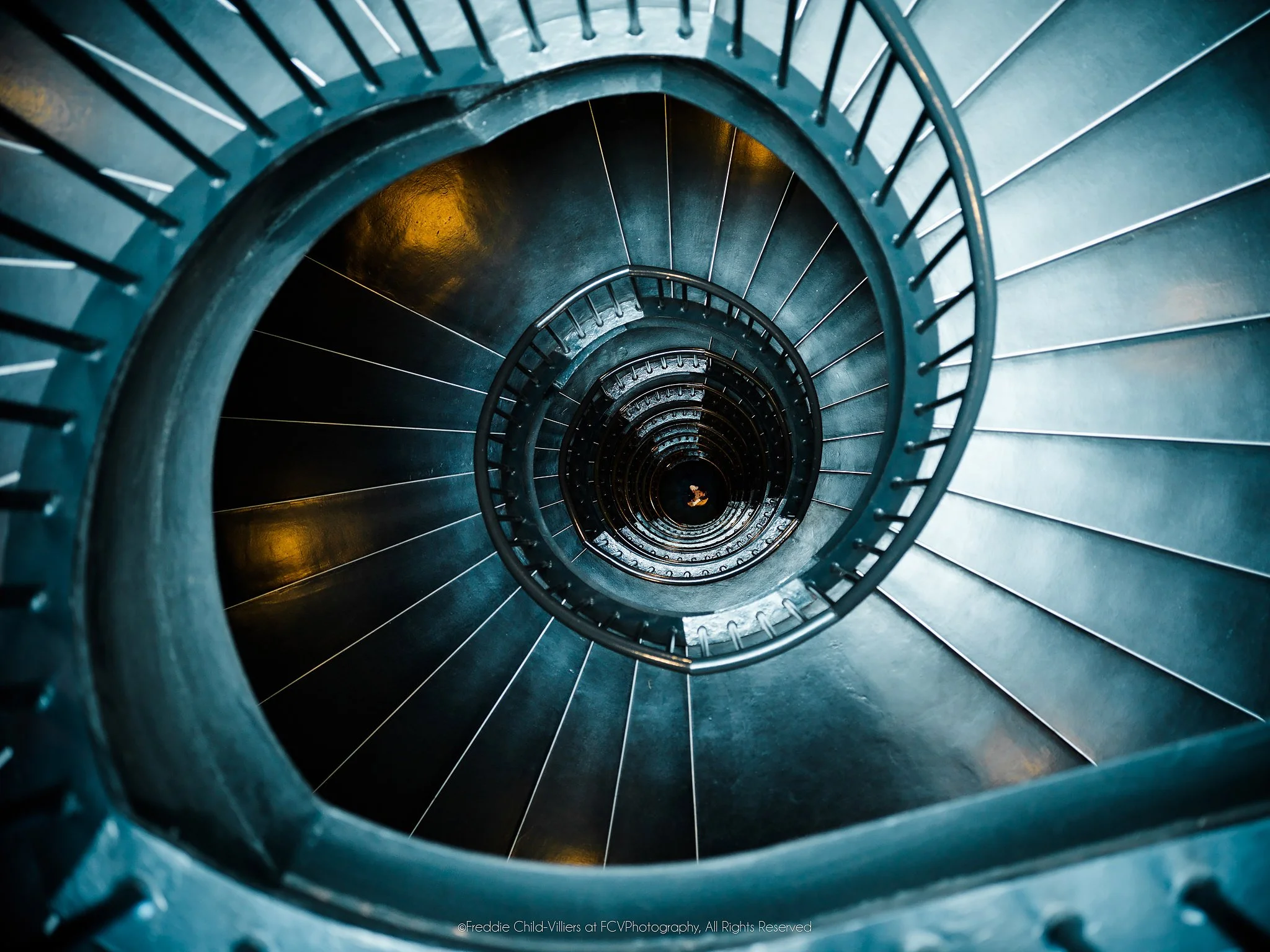 Teal-blue spiral staircase viewed from above, tightening around a deep central void with small amber lights.