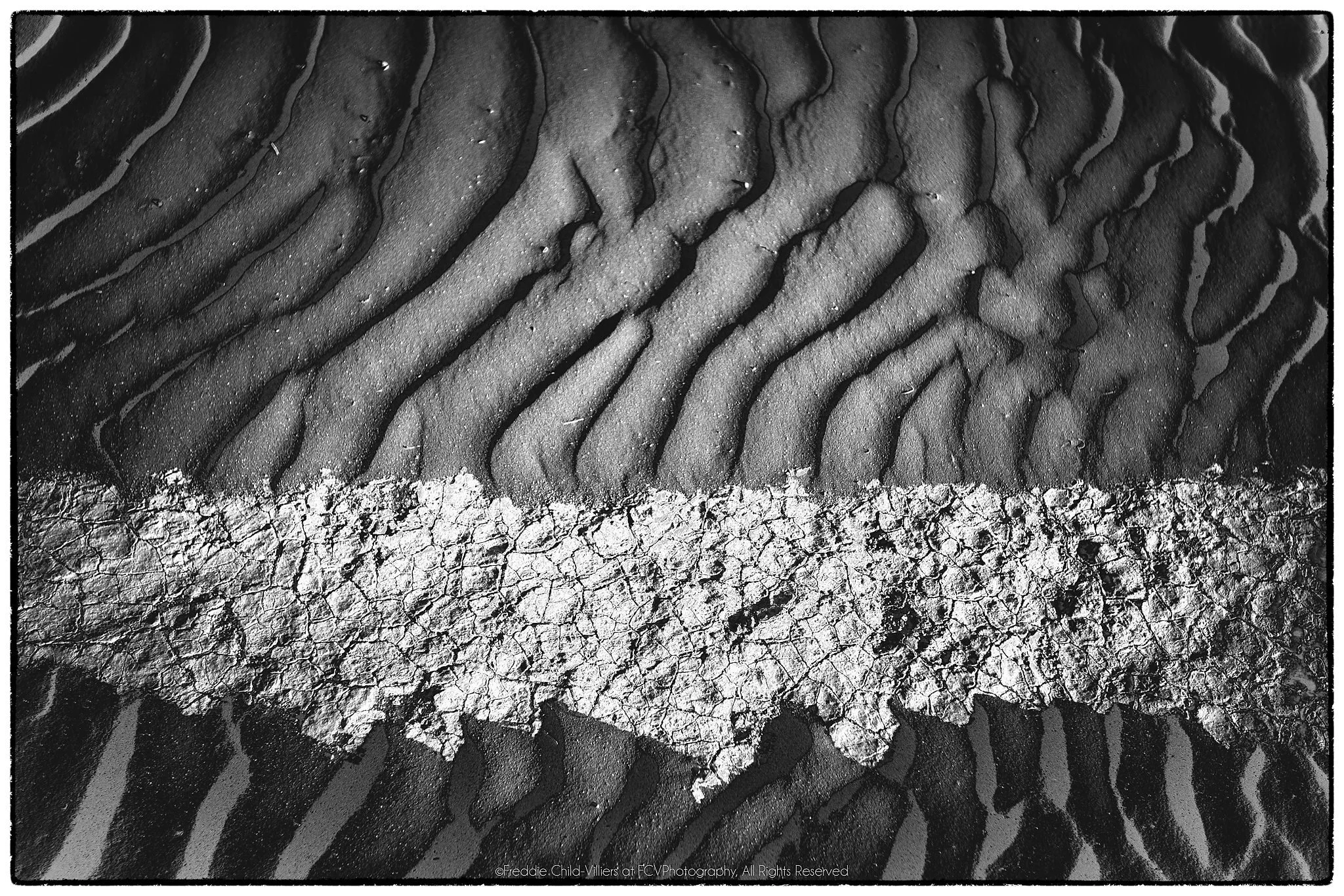 Pale cracked band crossing dark rippled sand in a high-contrast horizontal composition.