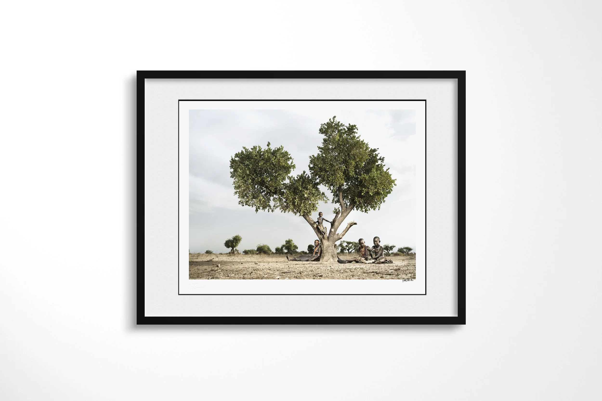 Framed presentation of Kenina & her sisters in a black frame and white mount, showing four Arbore sisters gathered beneath a broad tree, with one child perched in its branches.