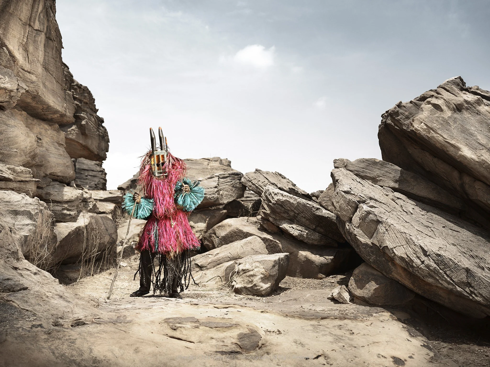 Walu (Mask), a Dogon masked figure in bright fibre costume standing in a narrow rocky passage.