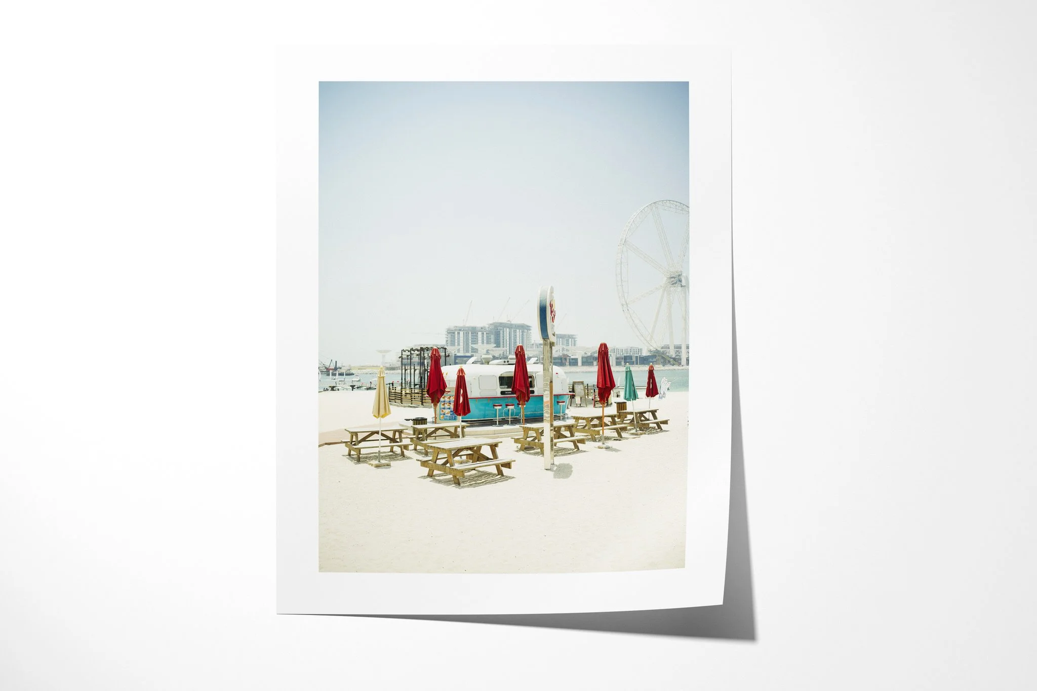 Fine art paper presentation of Desert Diner, depicting empty picnic tables and closed colourful parasols beside a small waterfront kiosk, with an observation wheel in the distance.