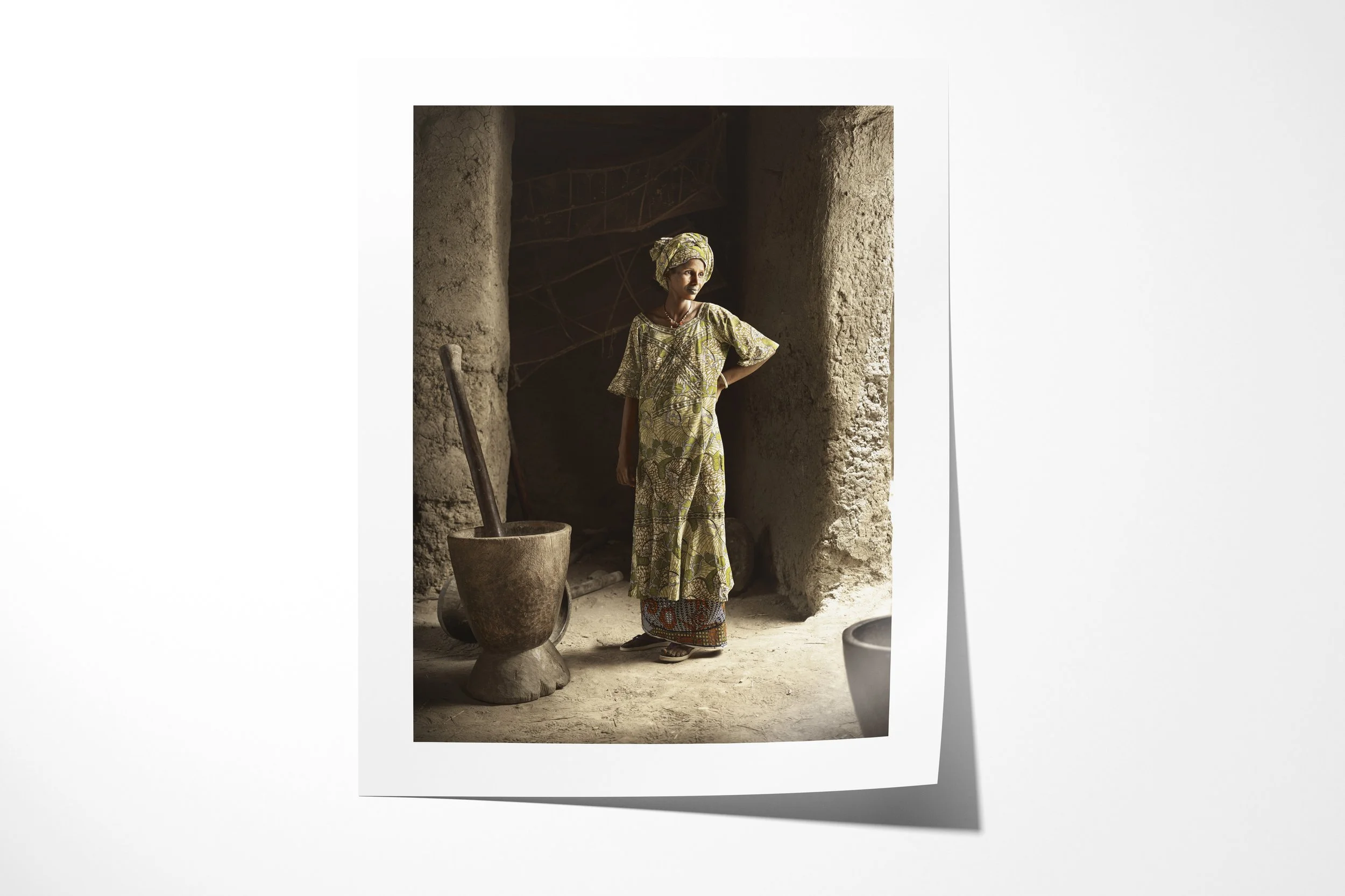 Fine art paper print of Tiedo, showing Tiedo standing beside a large wooden mortar in the doorway of a dim earthen building.