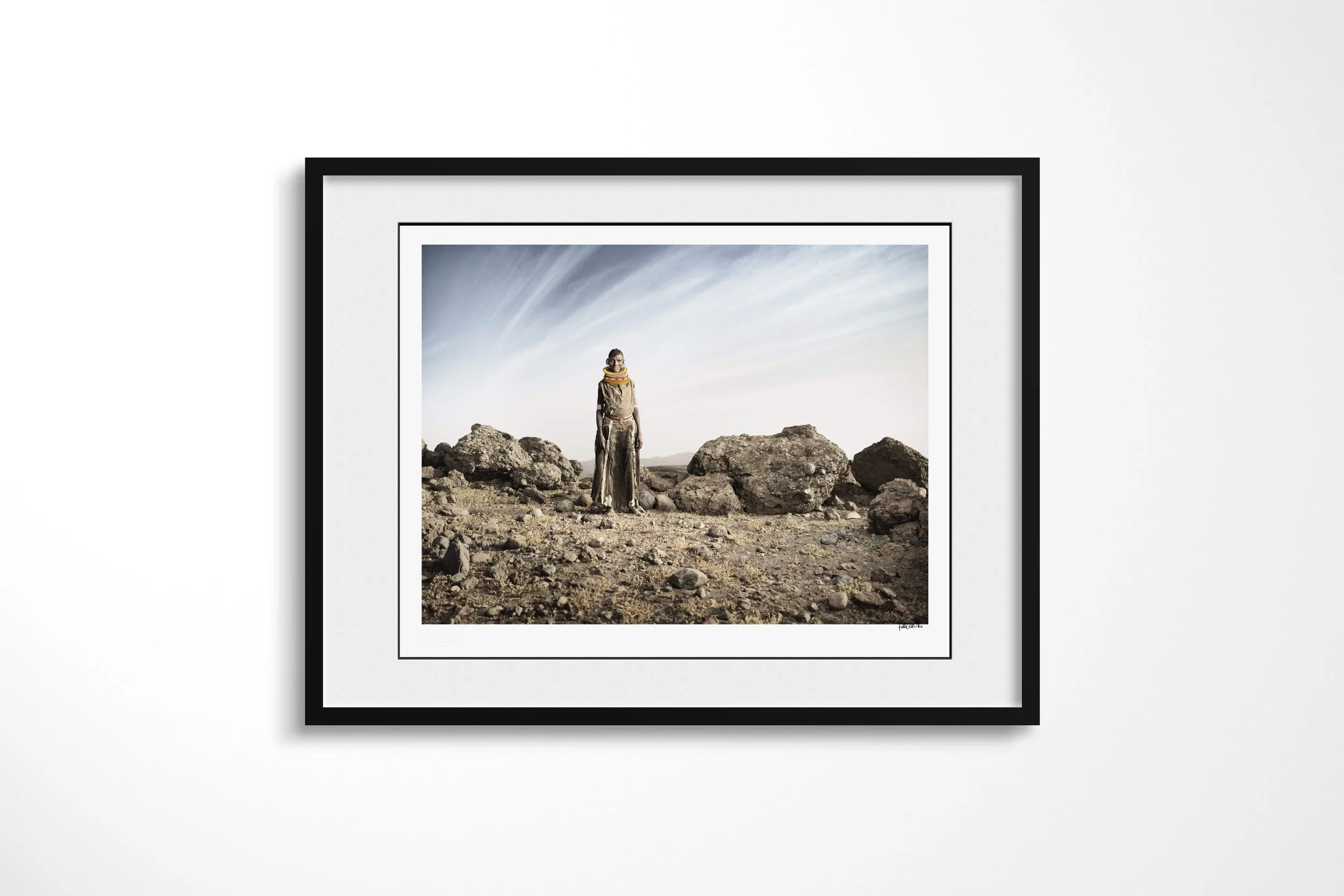 Framed presentation of Aseguan in a black frame and white mount, showing a Turkana woman in a long hide dress and beadwork standing among dark rocks in an arid landscape.
