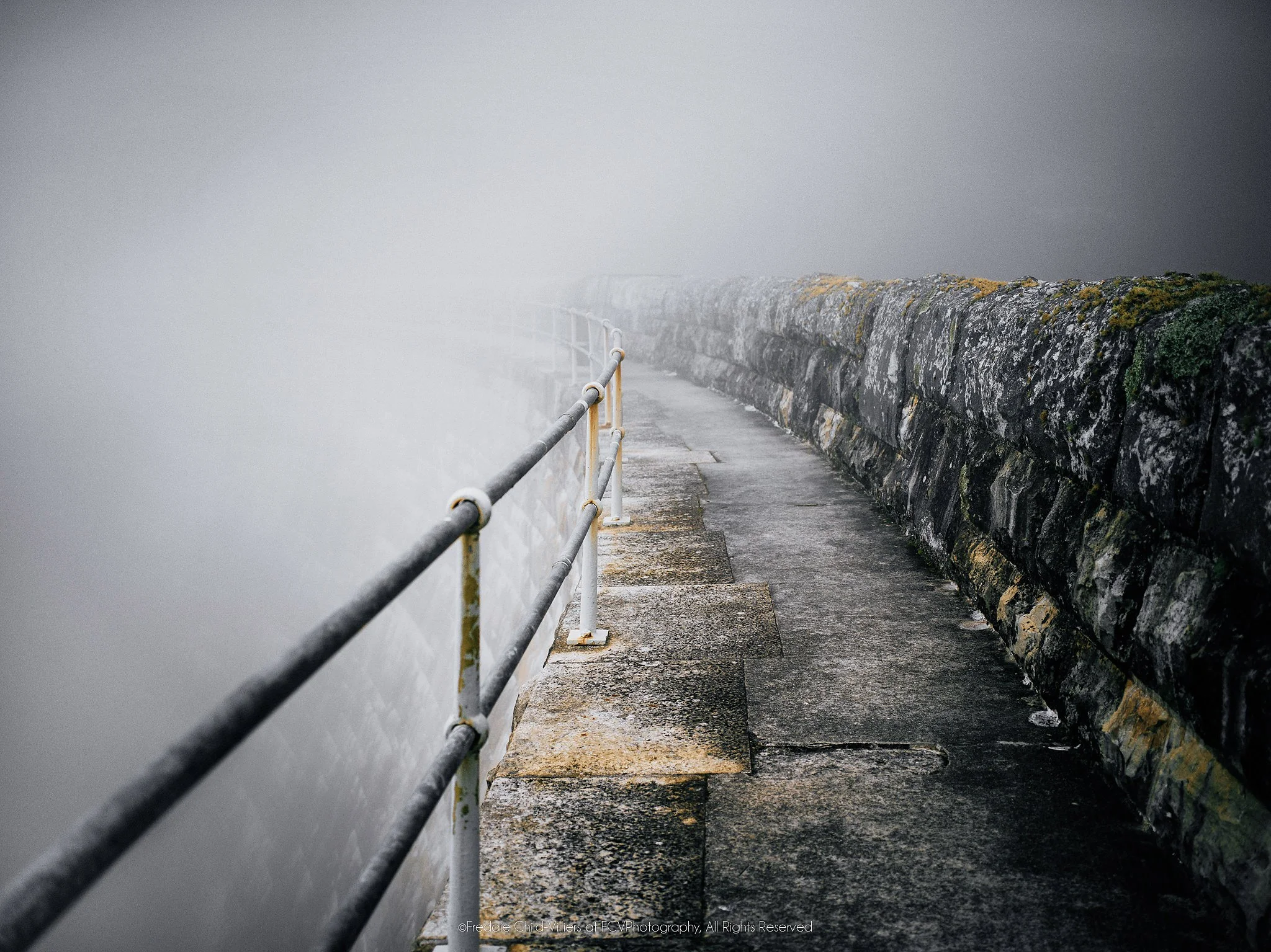 Weathered concrete walkway between a metal handrail and stone wall, curving out of sight into dense fog.