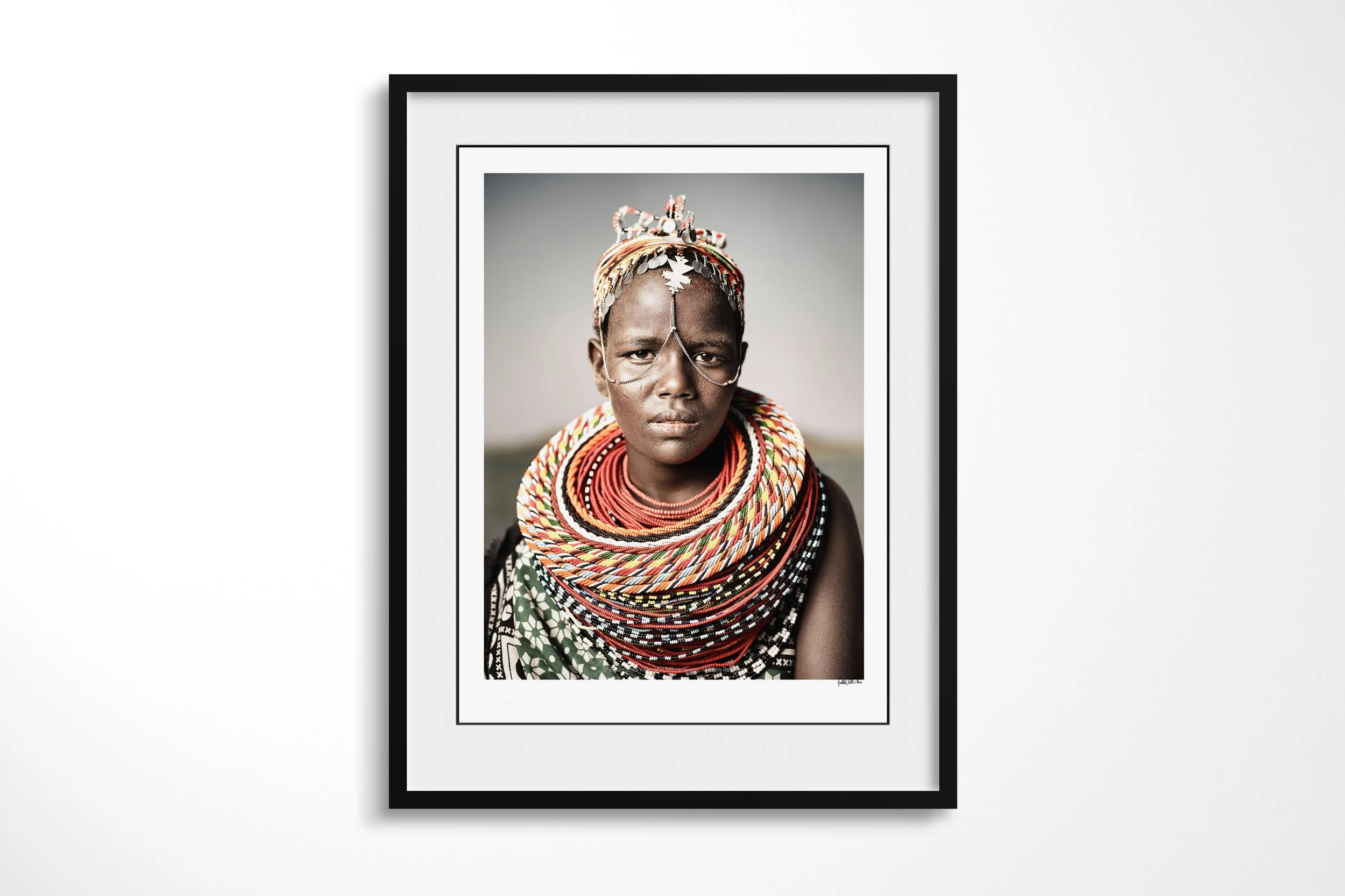 Framed presentation of Nadirie in a black frame and white mount, showing a close portrait of an El Molo woman wearing layered beadwork and fine chains across her face.