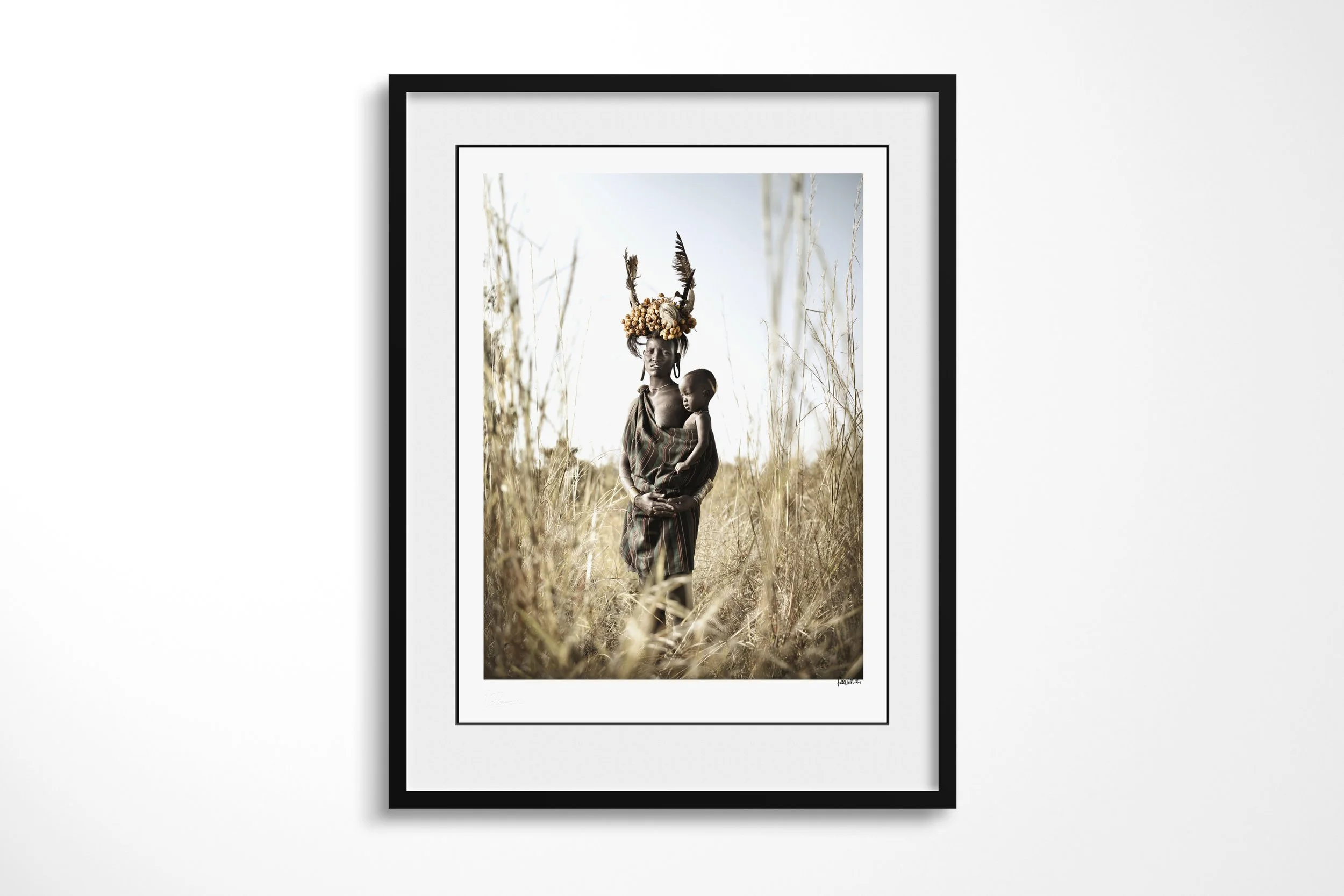 Framed presentation of Chomeri & Dhada in a black frame and white mount, showing a Mursi woman holding a child while wearing an elaborate headdress of horns, feathers and fruit.