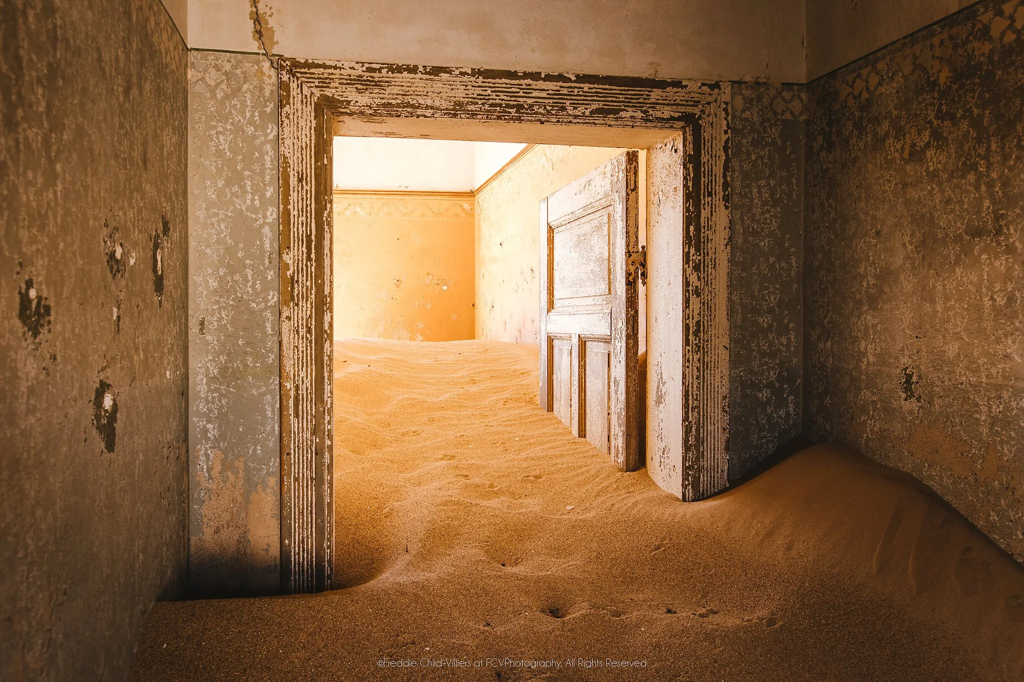Open doorways leading through worn ochre rooms whose floors are covered by rippled desert sand.