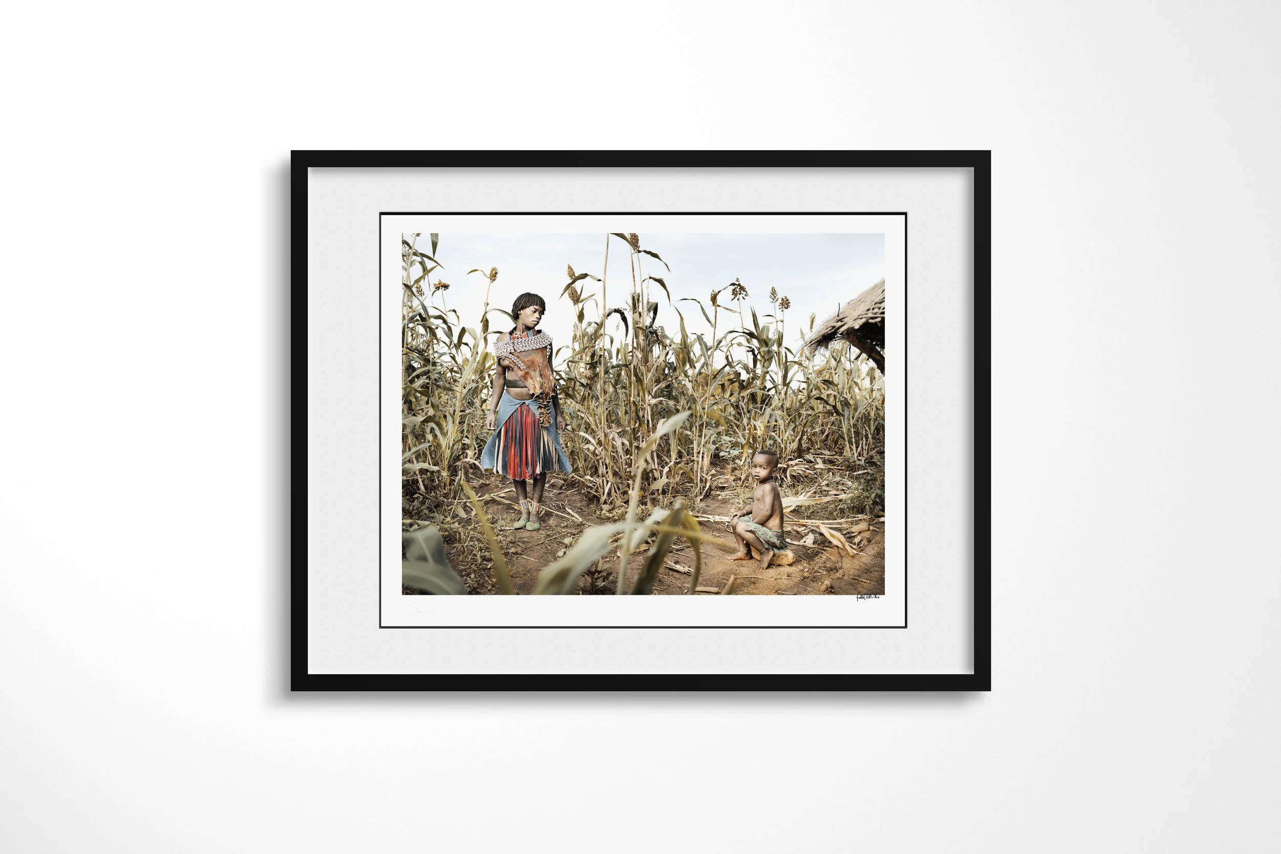 Framed presentation of Haila & Wancho in a black frame and white mount, showing a Banna woman and child standing among tall sorghum beside a thatched structure.