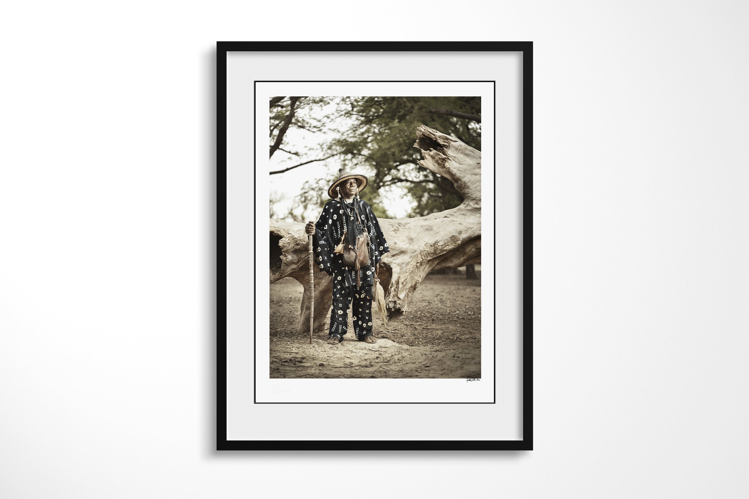 Framed presentation of Eli in a black frame and white mount, showing Eli in a patterned dark robe and broad hat holding a carved staff beneath a tree.