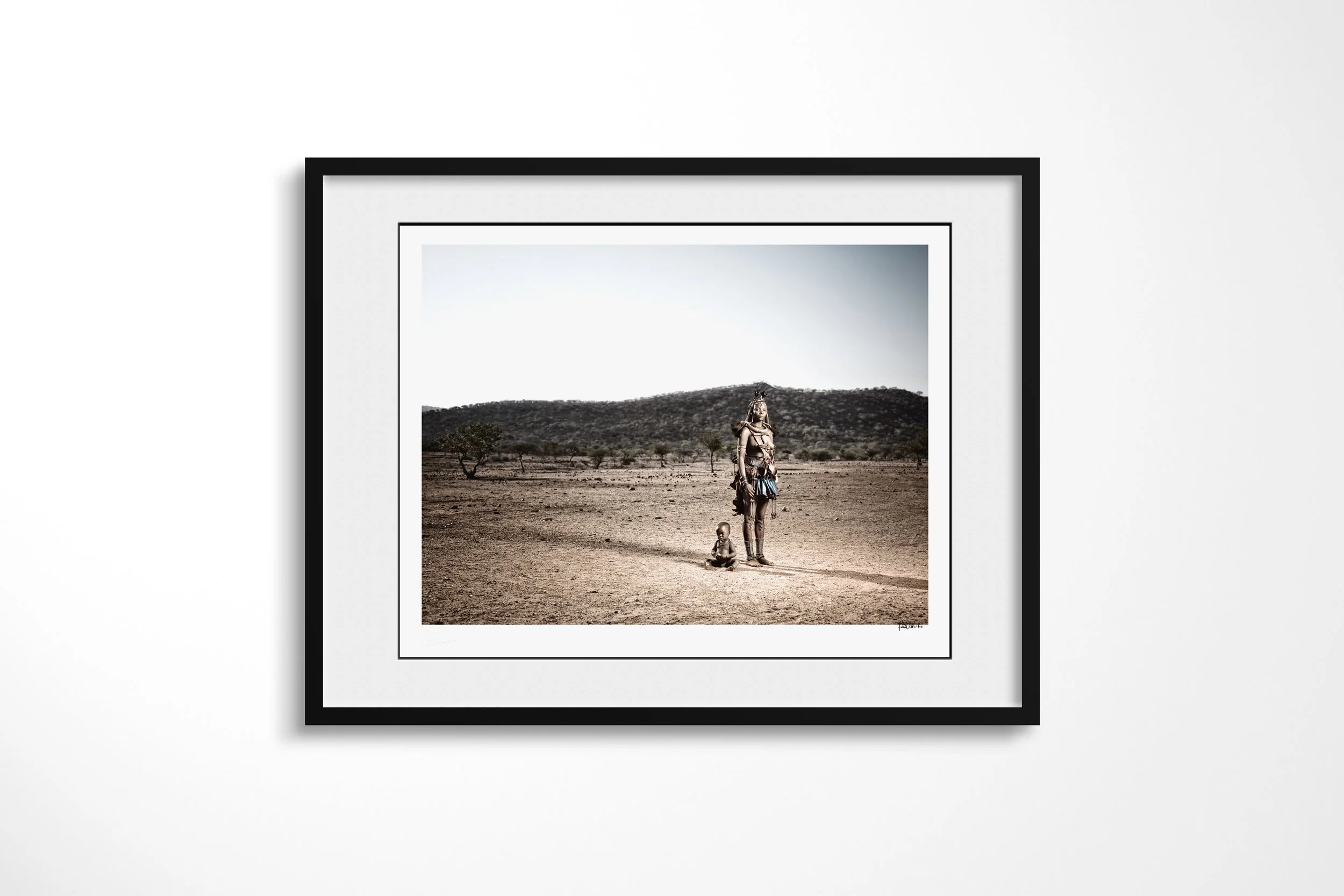 Framed presentation of Mburaivaka & Her Child in a black frame and white mount, showing Mburaivaka standing with her child in an expansive dry landscape backed by low hills.