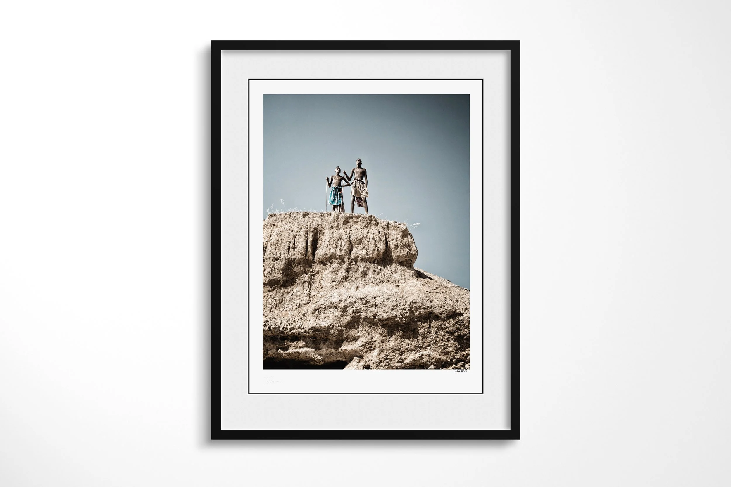 Framed presentation of Mbangu (left) and Turimuno (right) in a black frame and white mount, showing Mbangu and Turimuno standing together on the edge of a high eroded rock formation.