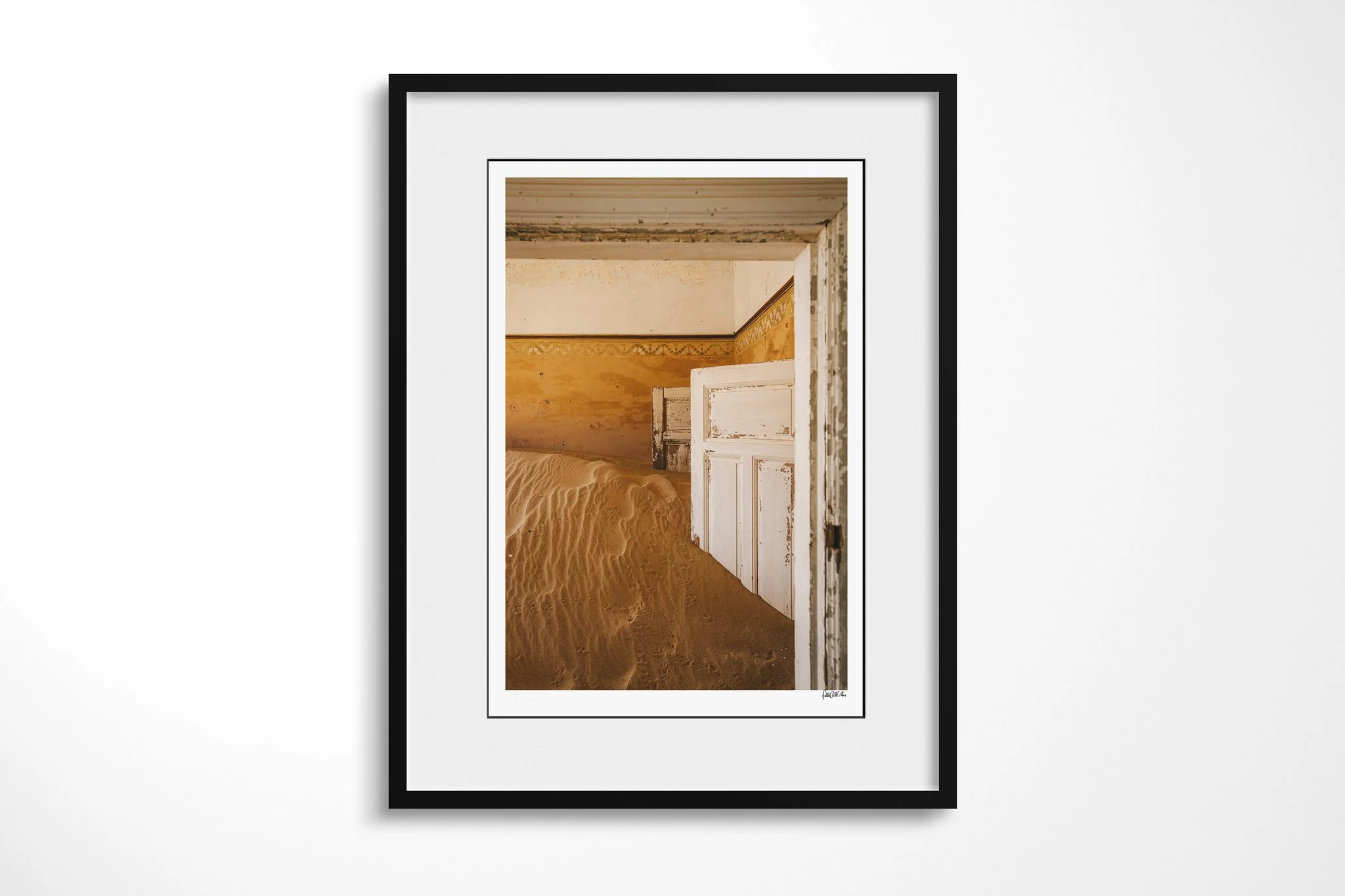 Black-framed fine art print of Doorway III with a white mount, showing chipped white doors and ochre walls layered behind a high drift of rippled sand in an abandoned interior.