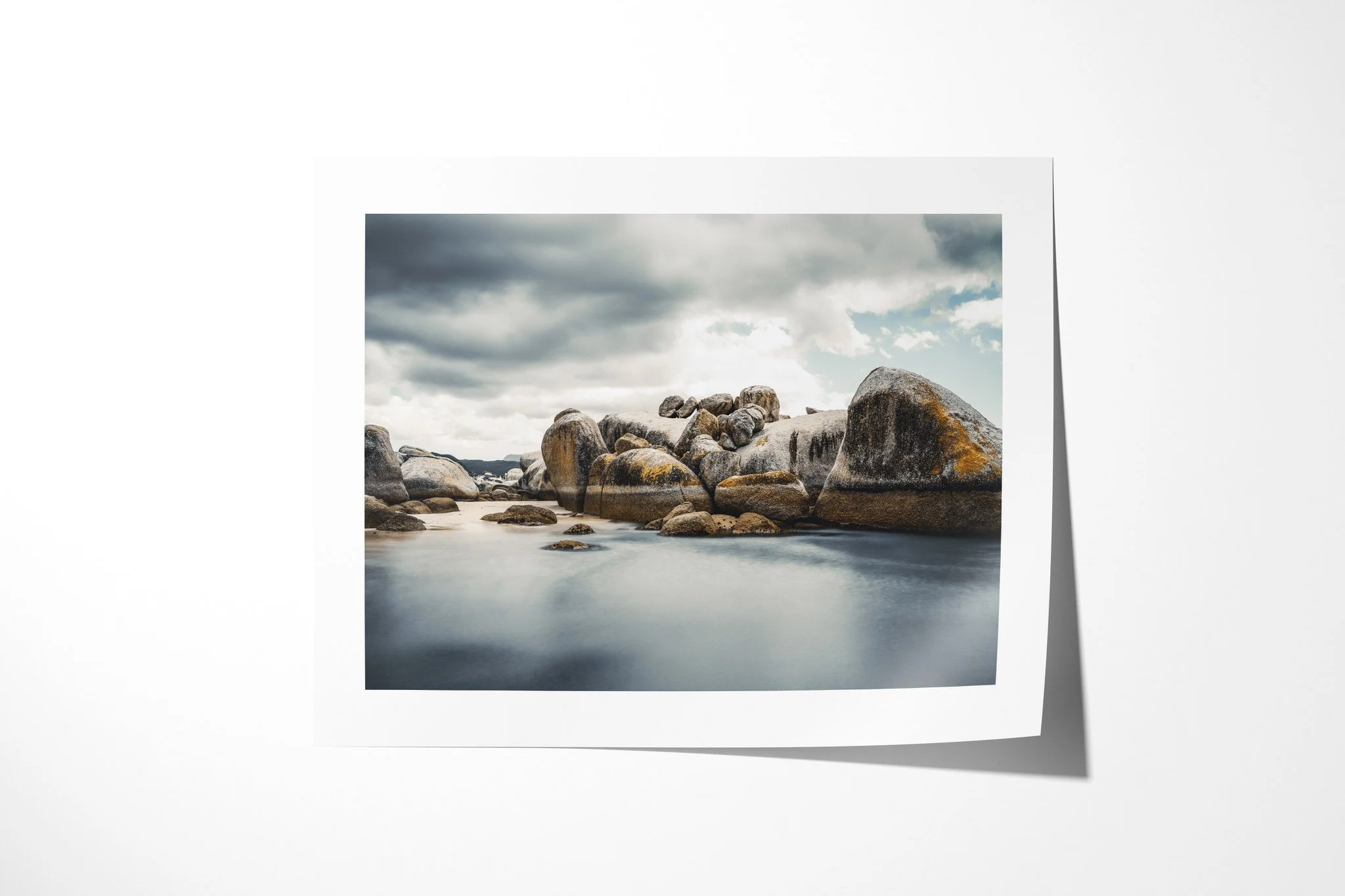 Fine art paper print of Tideline, showing rounded granite boulders around smooth blue-grey coastal water beneath heavy clouds, with patches of warm lichen.