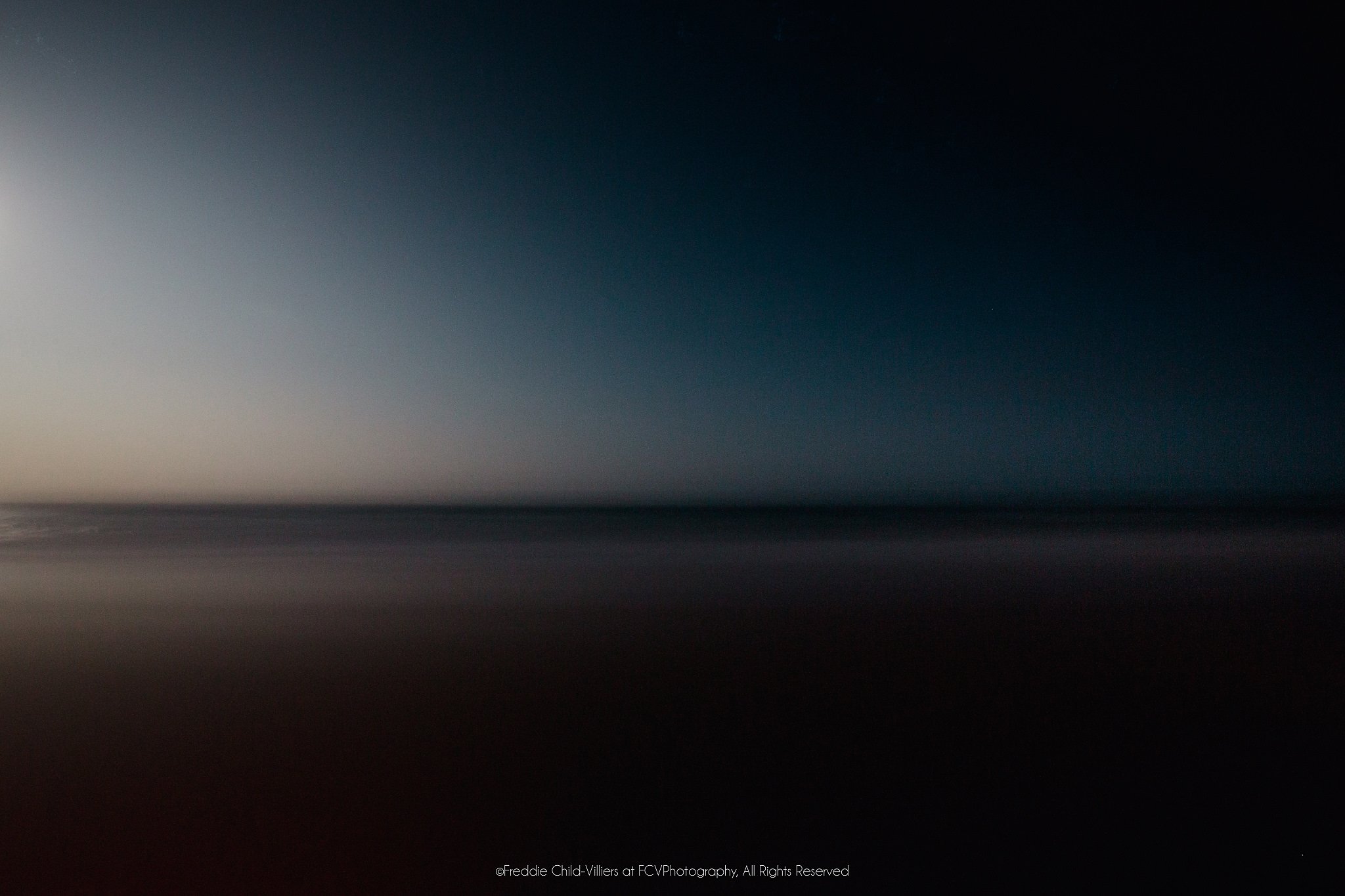 Soft, nearly abstract bands of dark blue sky, muted sea and reddish foreground divided by a faint horizon.