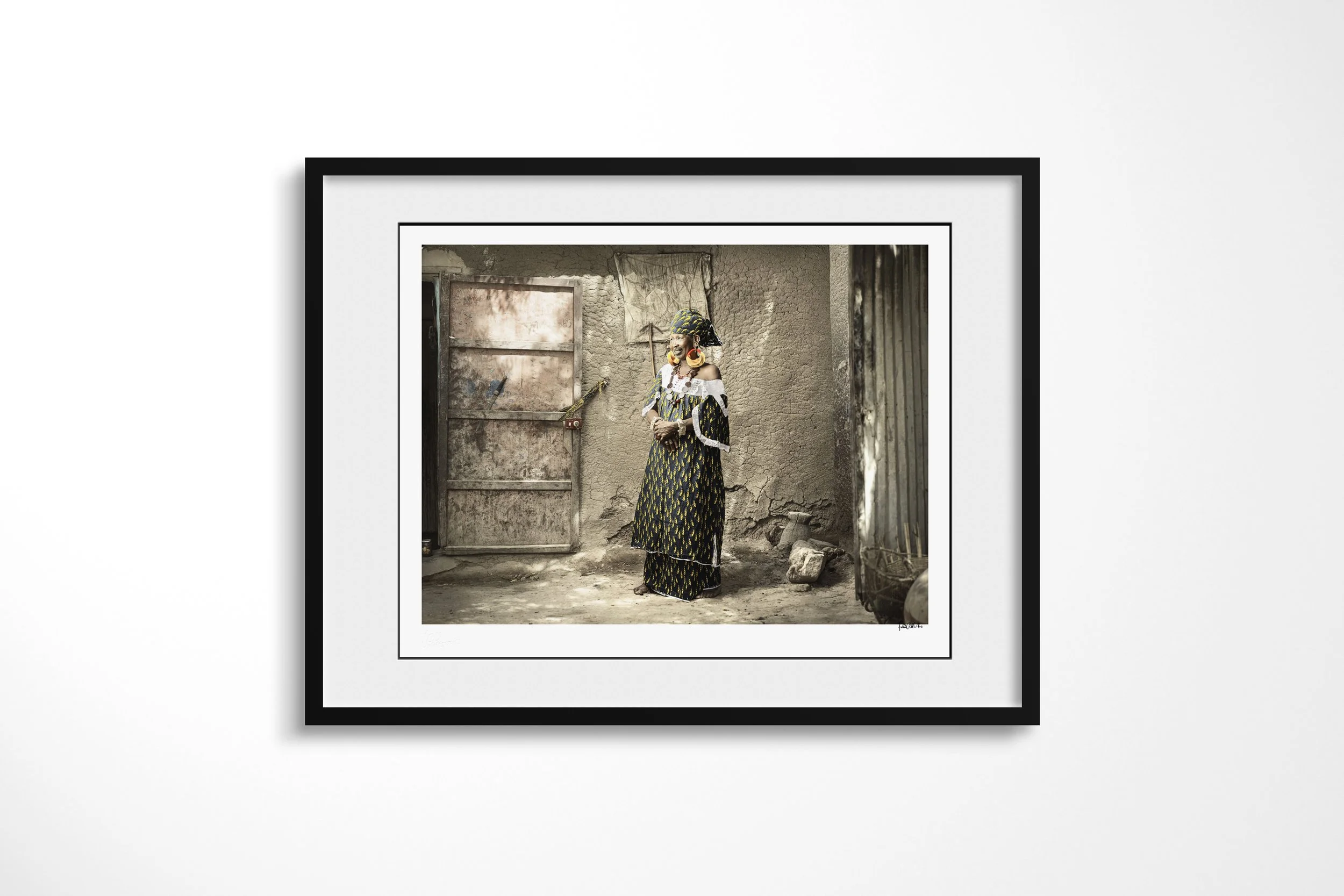 Framed presentation of Thindel in a black frame and white mount, showing Thindel wearing large gold earrings and patterned clothing beside a weathered metal door.