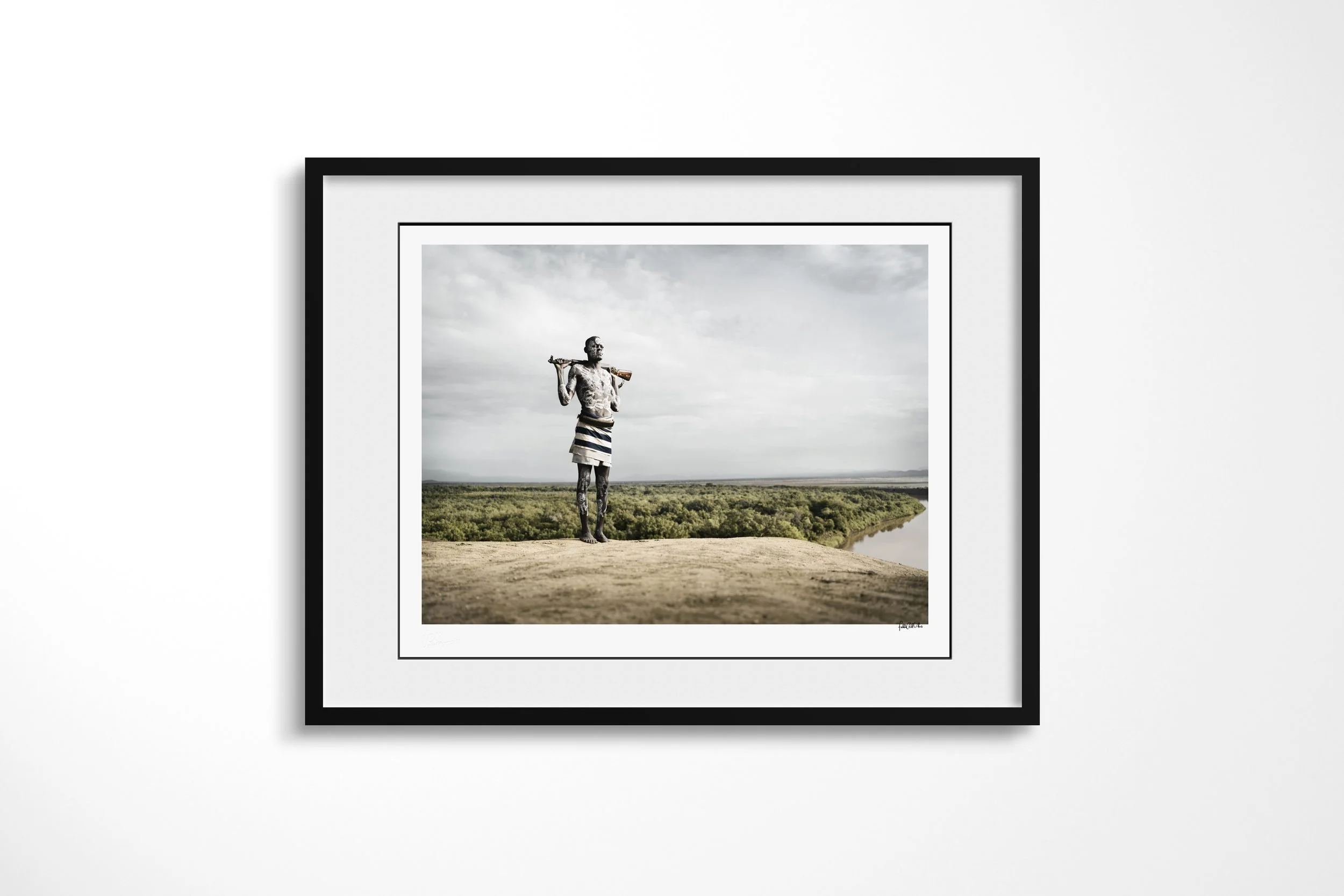 Framed presentation of Muda in a black frame and white mount, showing a Kara man with painted skin and a rifle across his shoulders overlooking the Omo landscape.