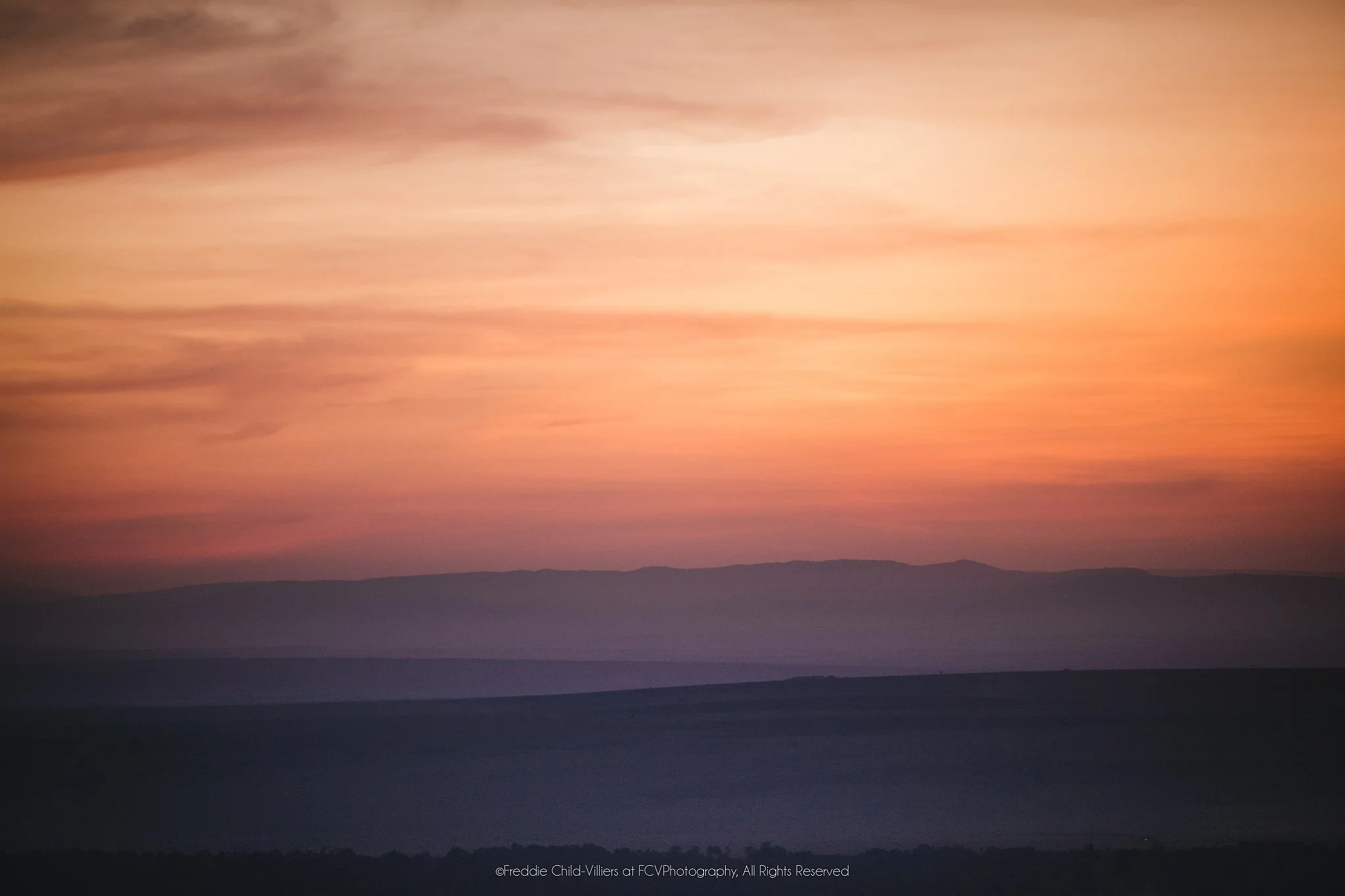 Soft horizontal bands of salmon, orange and mauve above blurred dark hills at sunrise.