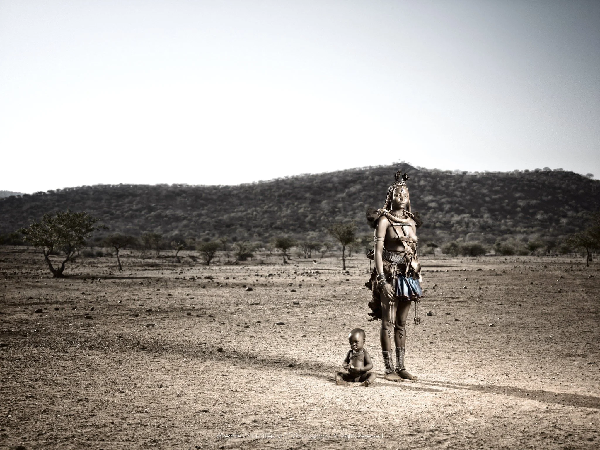 Mburaivaka standing with her child in an expansive dry landscape backed by low hills.