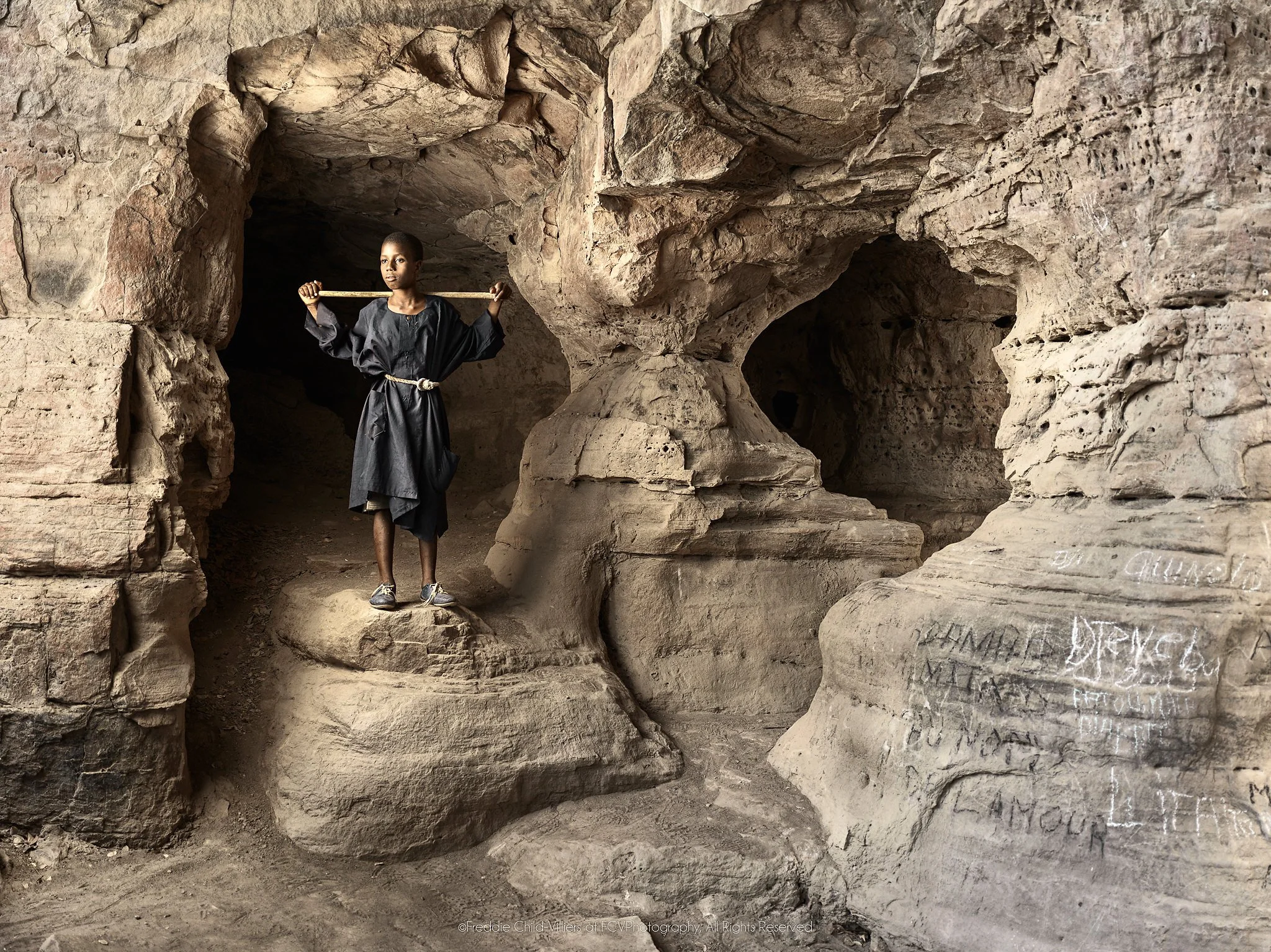 Modibo standing with outstretched arms inside a cave marked with handwritten requests.