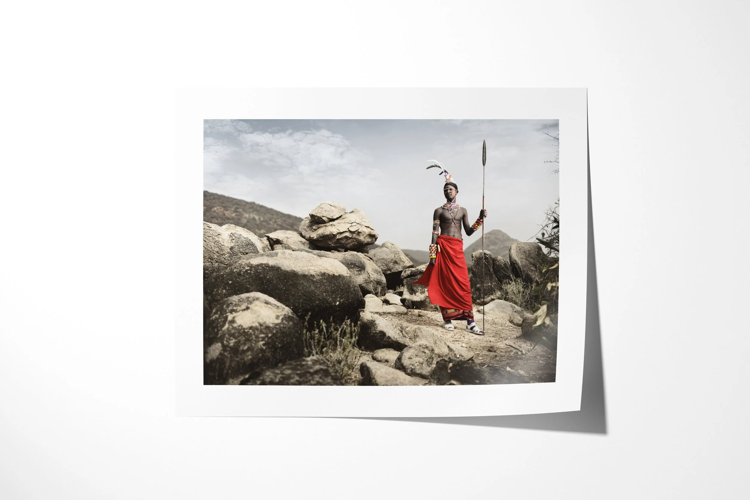 Fine art paper print of Lempati, showing a Samburu warrior in a red wrap holding a spear among large rocks and mountain scrub.