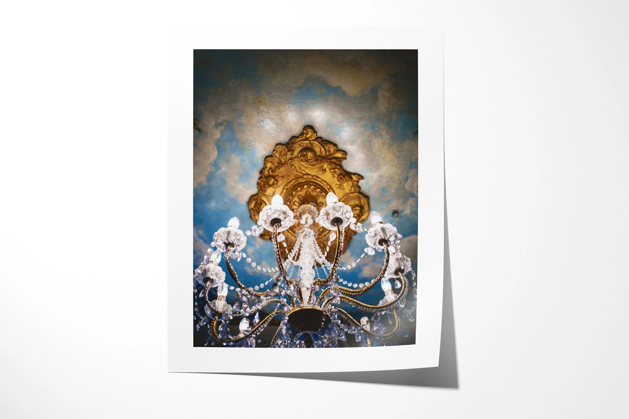 Chandelier presented as an unframed fine art paper print, depicting an ornate crystal chandelier viewed from below against a gilded ceiling rose and cracked blue plaster.
