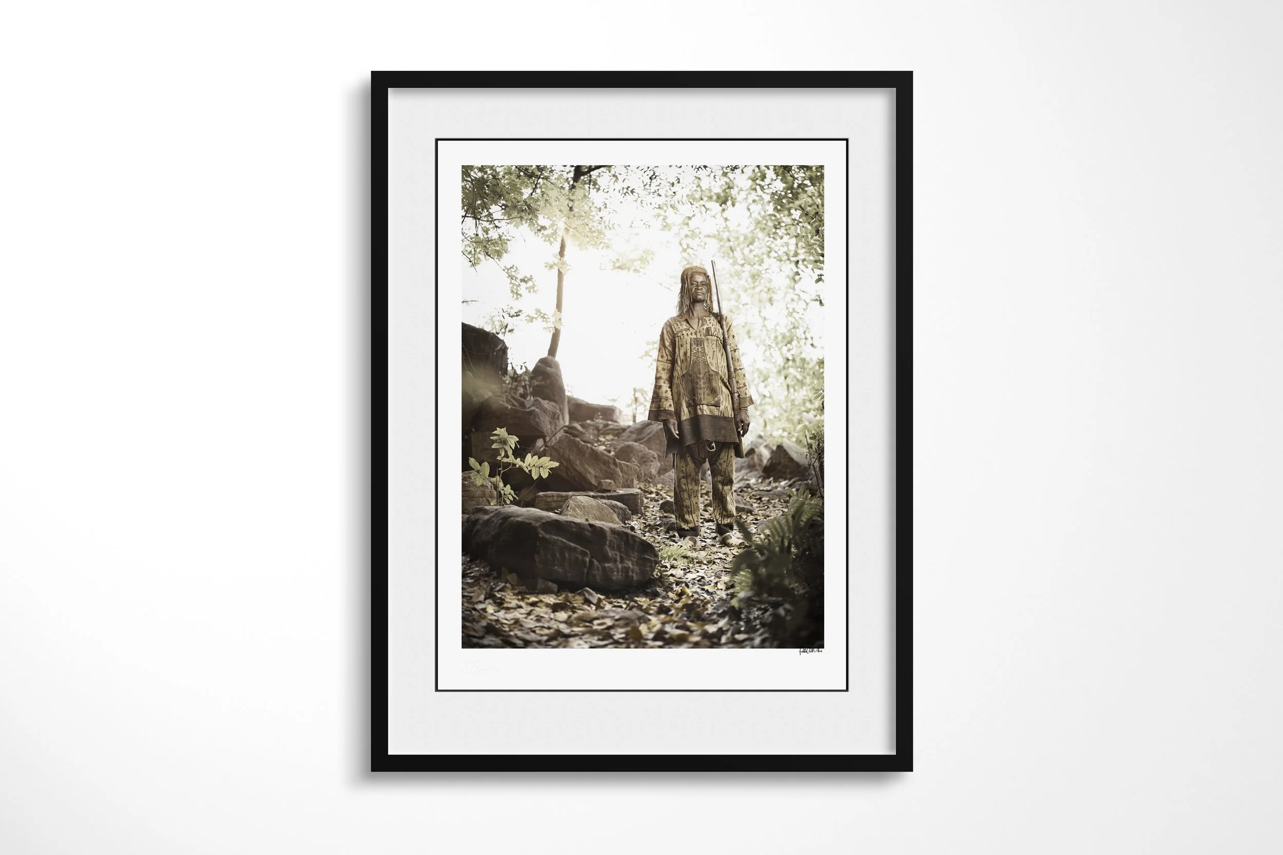 Framed presentation of Bandjougou in a black frame and white mount, showing Bandjougou standing among rocks and trees in a shaded green valley.