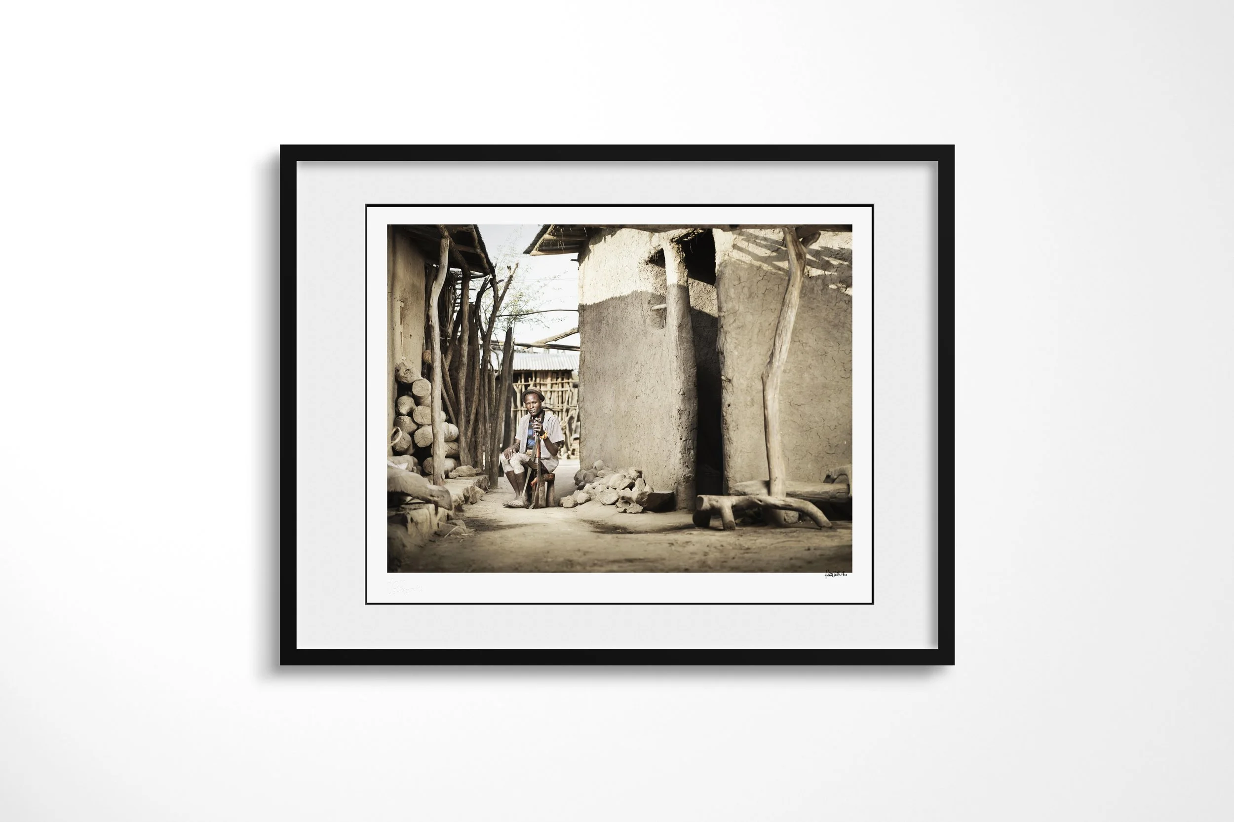 Framed presentation of Bule in a black frame and white mount, showing an Arbore man seated between earthen buildings with a staff and pale-painted legs.