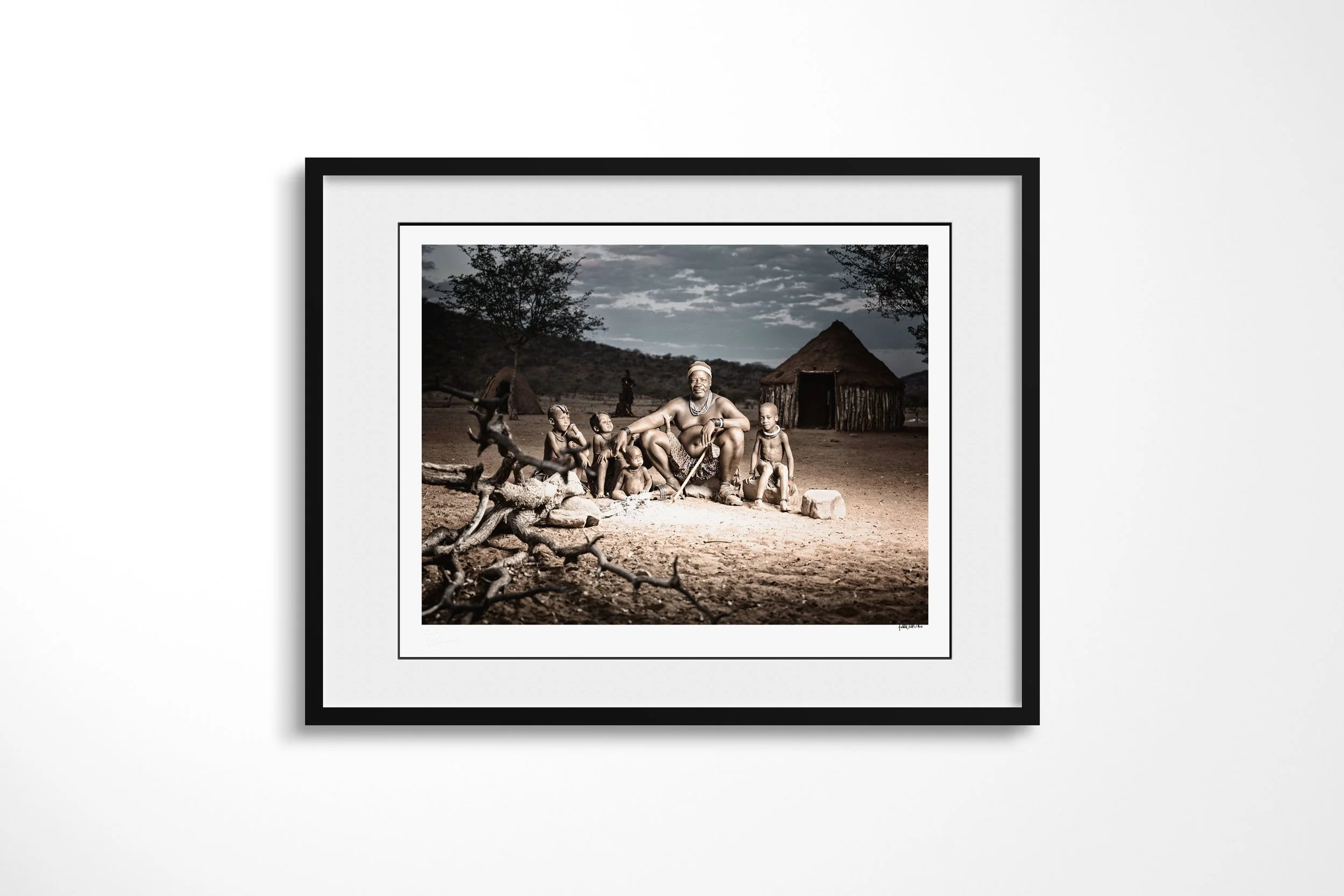 Framed presentation of Chief Tjikoko in a black frame and white mount, showing Chief Tjikoko seated with children beside the ancestral fire outside a conical Himba dwelling.