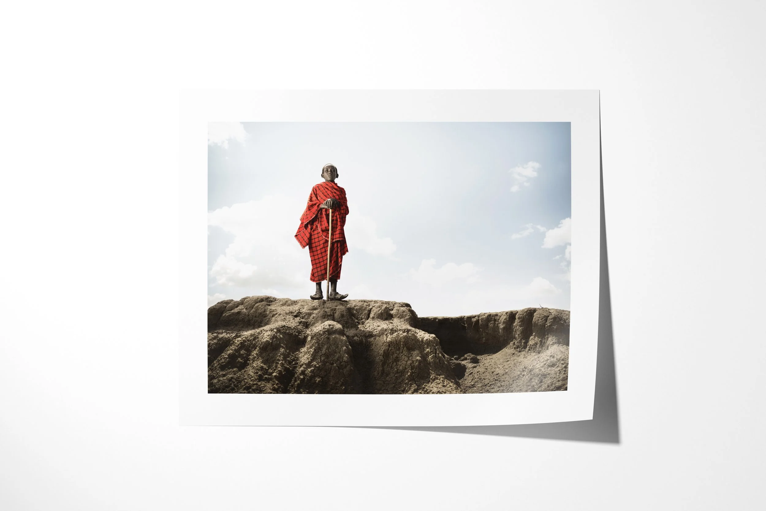 Fine art paper print of Saitoti, showing a Maasai boy in a red checked wrap standing with a herding staff on an eroded ridge.