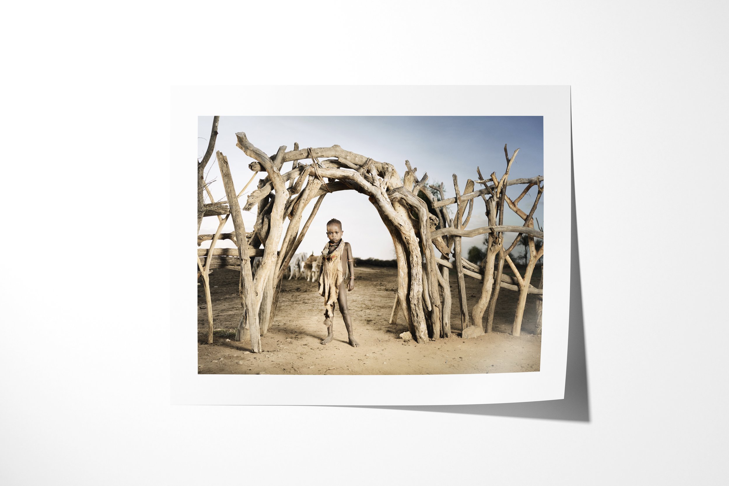 Fine art paper print of Gadi, showing a young Hamar boy standing beneath a curved entrance of interwoven branches in a dry homestead.