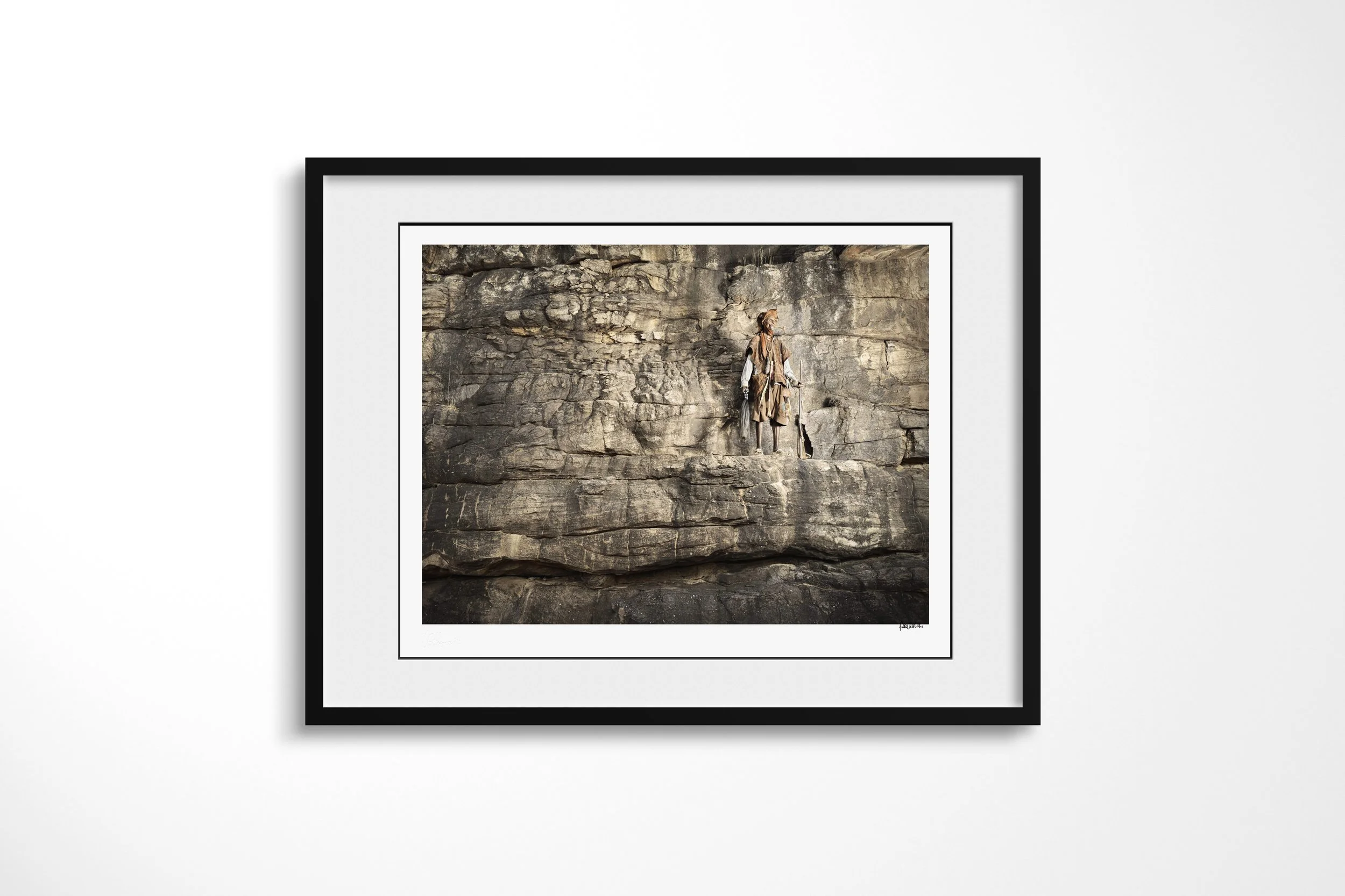 Framed presentation of Anbaga in a black frame and white mount, showing Anbaga standing alone against a vast banded sandstone cliff.