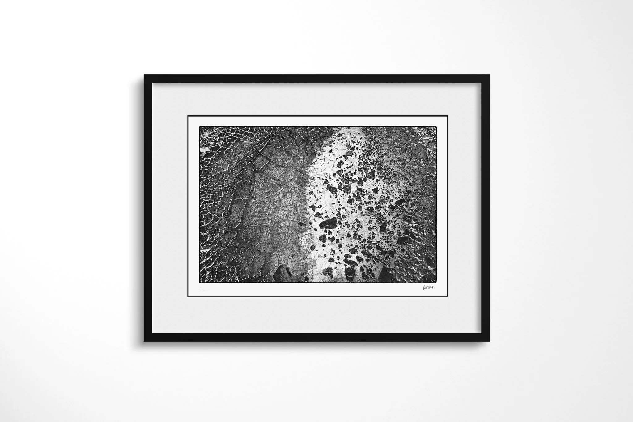 Framed presentation of Chalbi Desert in a black frame with a wide white mount, showing cracked earth in black and white, divided by a broad pale curving band scattered with dark stones.