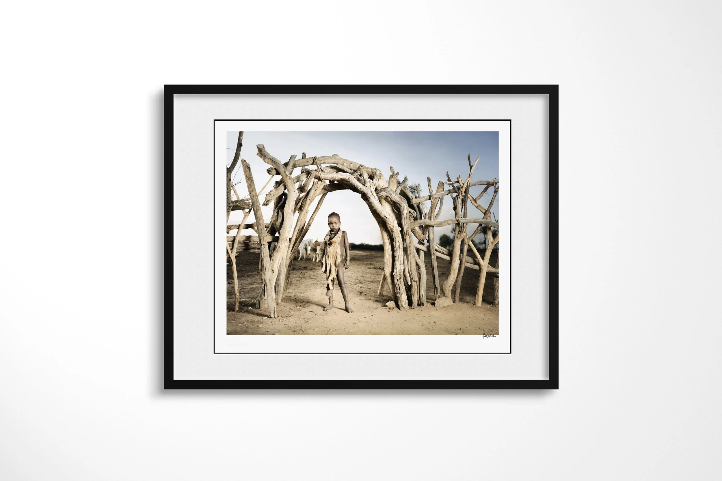 Framed presentation of Gadi in a black frame and white mount, showing a young Hamar boy standing beneath a curved entrance of interwoven branches in a dry homestead.